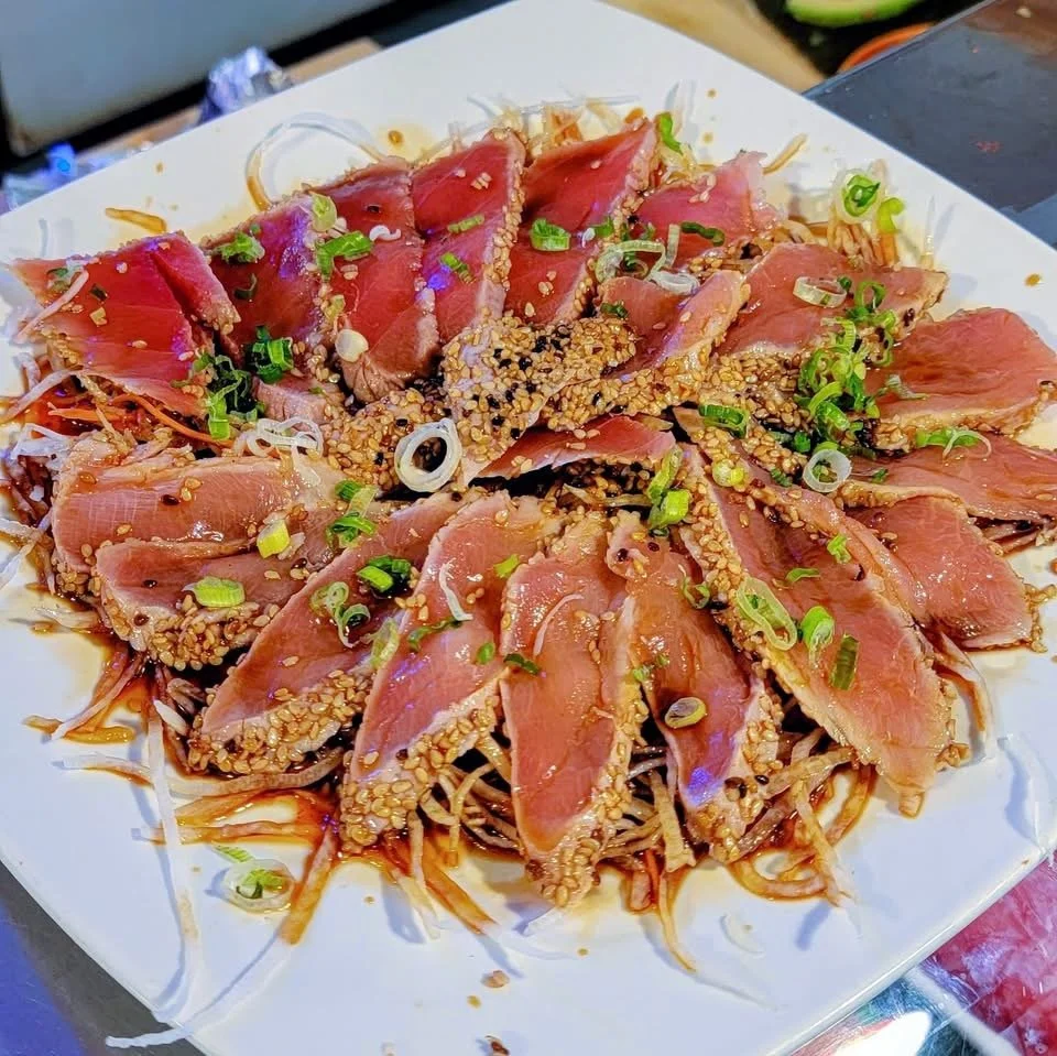 Sliced seared tuna with sesame seeds and chopped green onions on a bed of shredded vegetables and noodles, garnished with a sauce or dressing on a white plate.