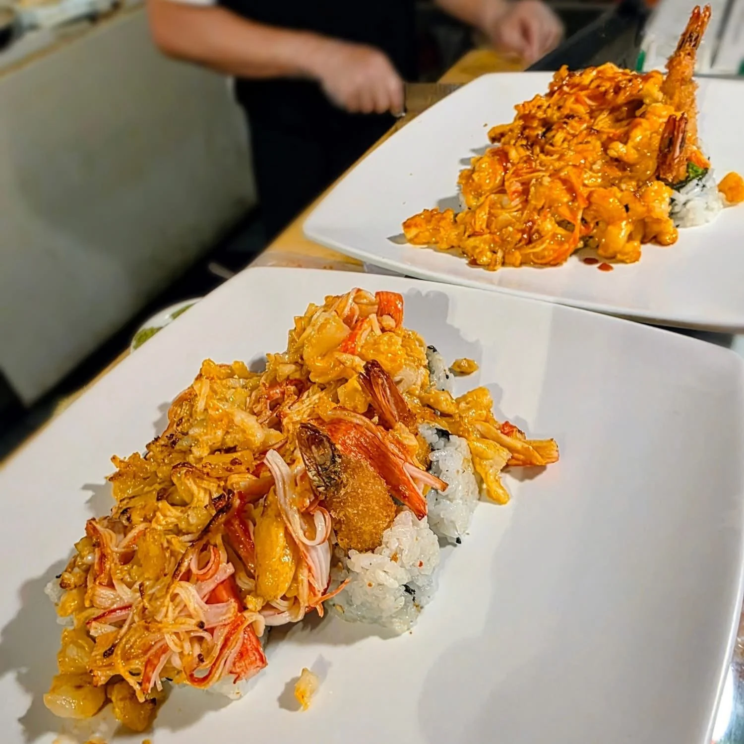 Close-up of a sushi roll topped with shrimp, crab, and a spicy orange sauce, served on a white plate.