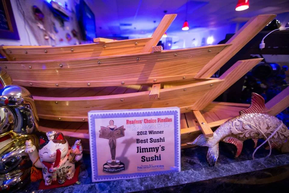 A wooden boat model on display with a sign indicating it was awarded the 2012 Readers' Choice Pinellas Best Sushi at Jimmy's Sushi. There are decorative items including a Maneki-neko figurine and a fish sculpture nearby.