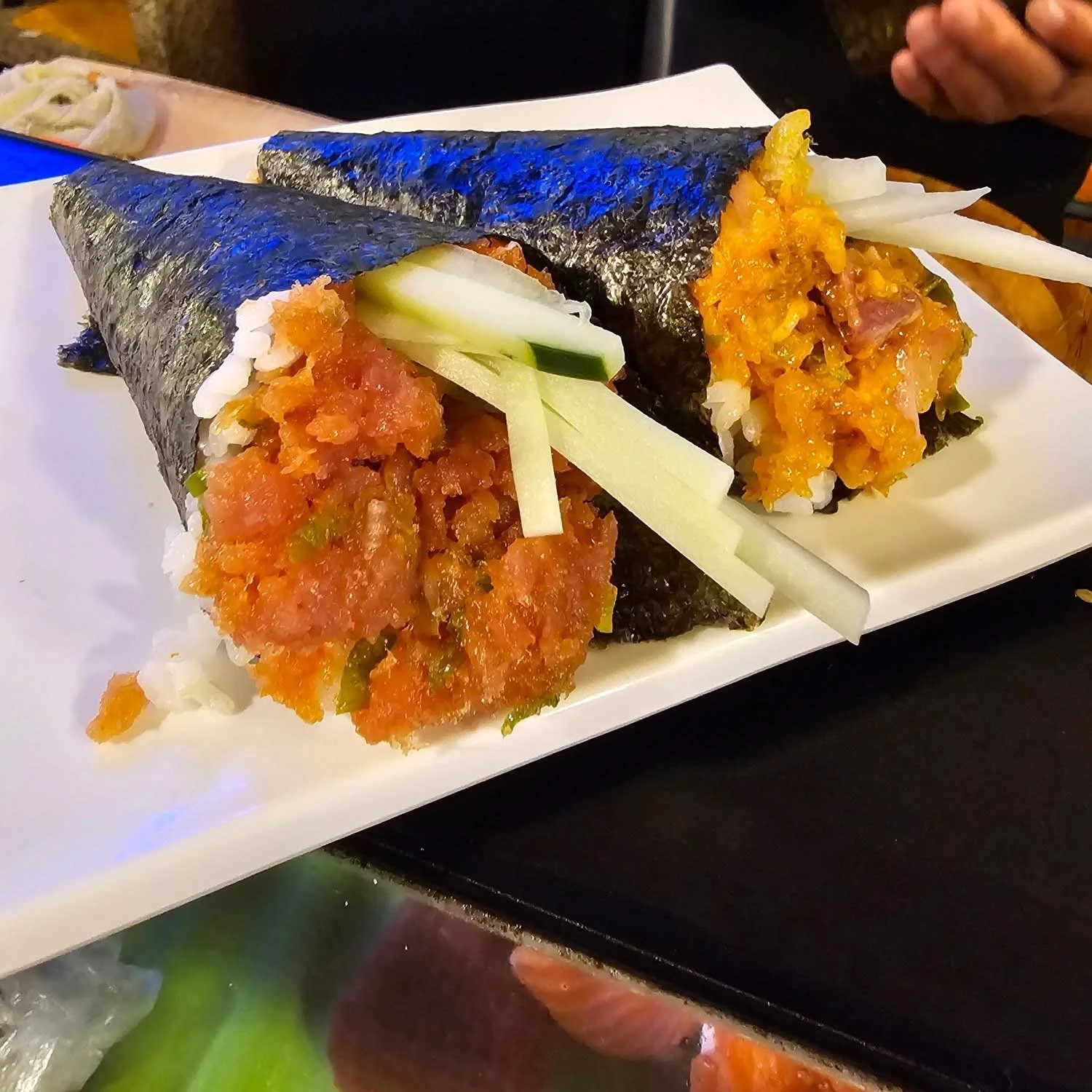 Two sushi hand rolls with nori seaweed, filled with rice, spicy tuna, cucumber, and other vegetables, served on a white rectangular dish.