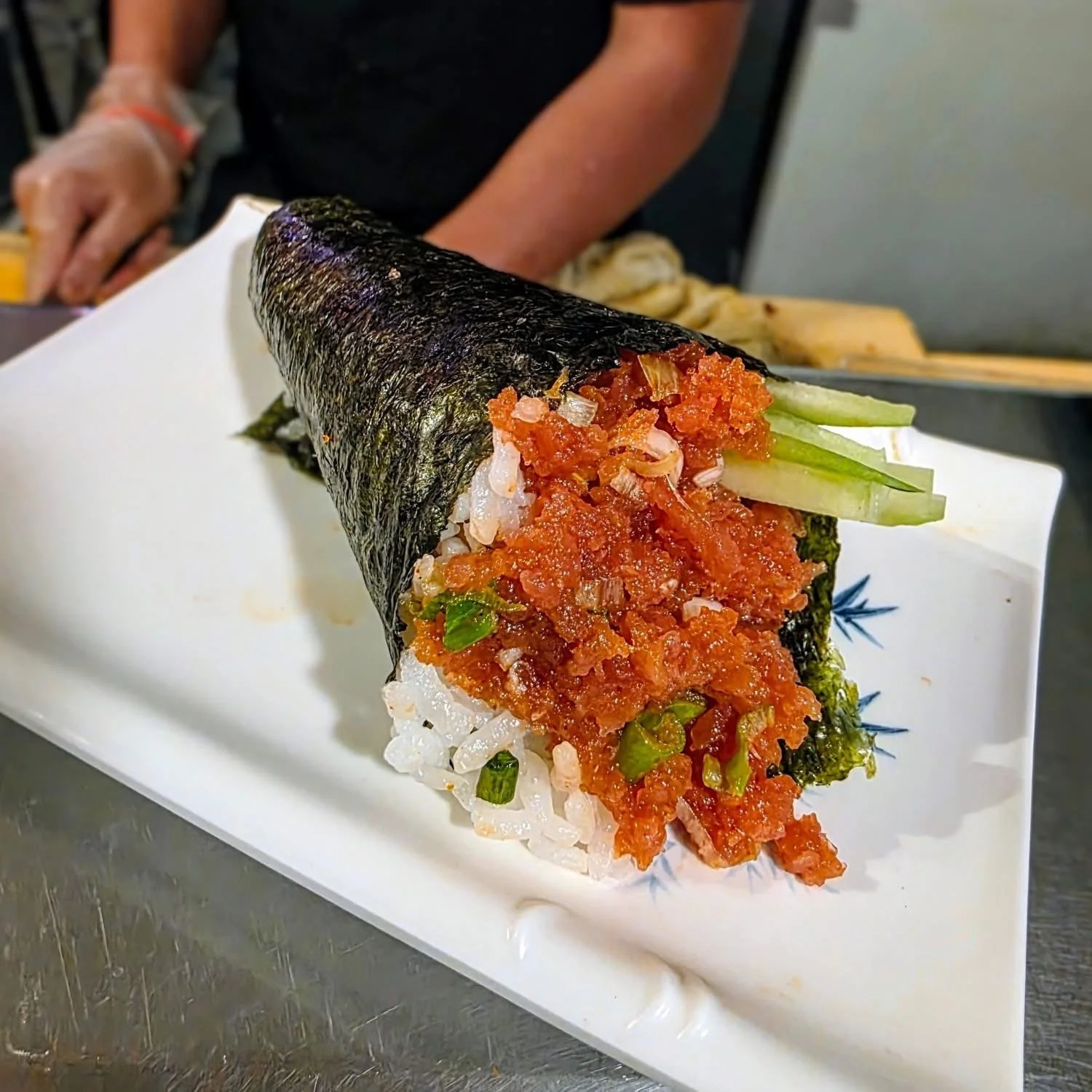 A large hand roll sushi with nori seaweed wrapping, filled with rice, spicy tuna, cucumber, and green onion, served on a white plate.