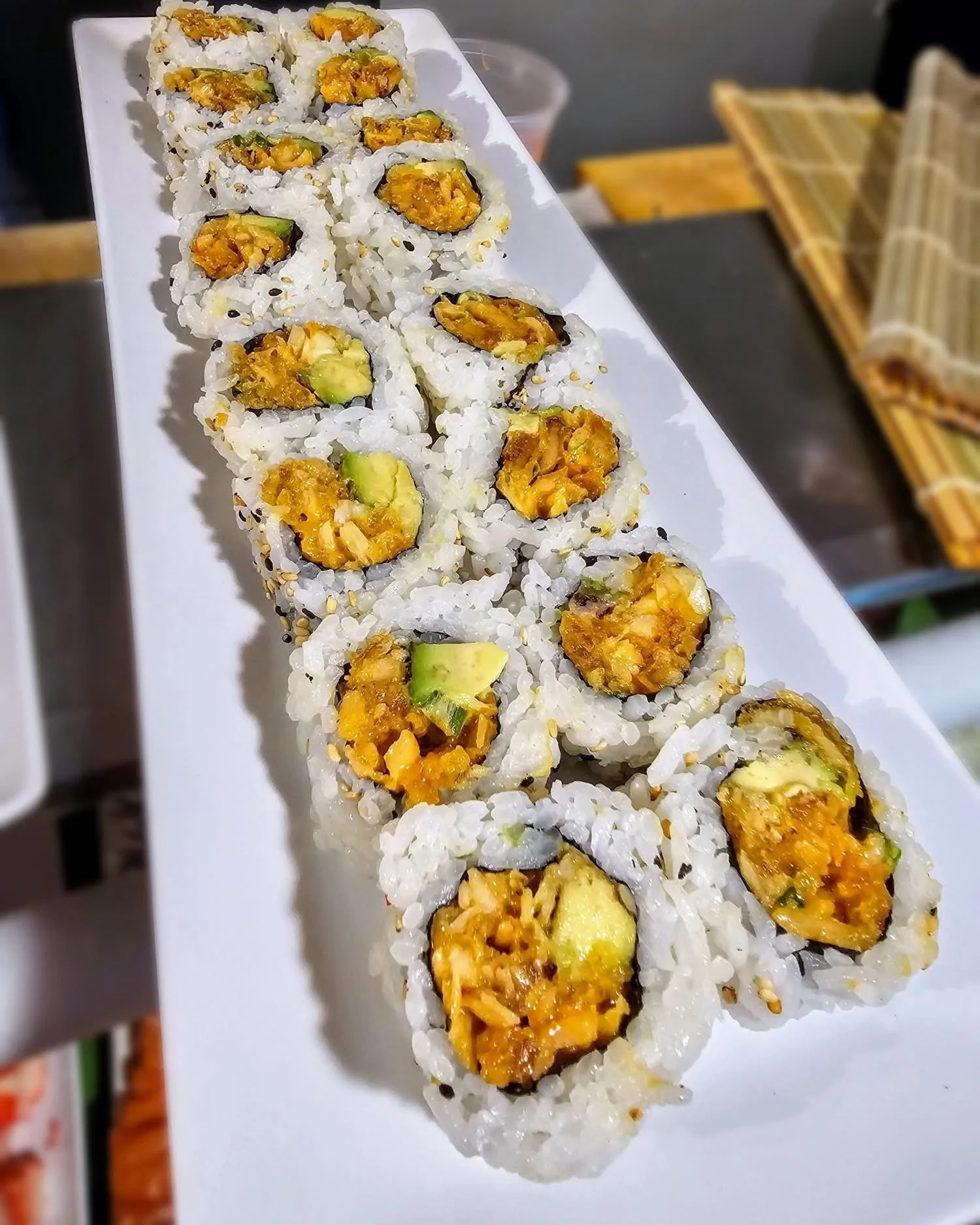 A white rectangular platter of sushi rolls filled with avocado and spicy filling, topped with sesame seeds.