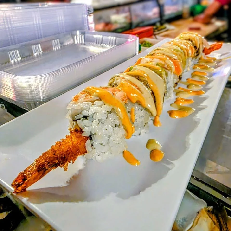 A long sushi roll with shrimp tempura, topped with orange and green sauces, on a white rectangular plate.