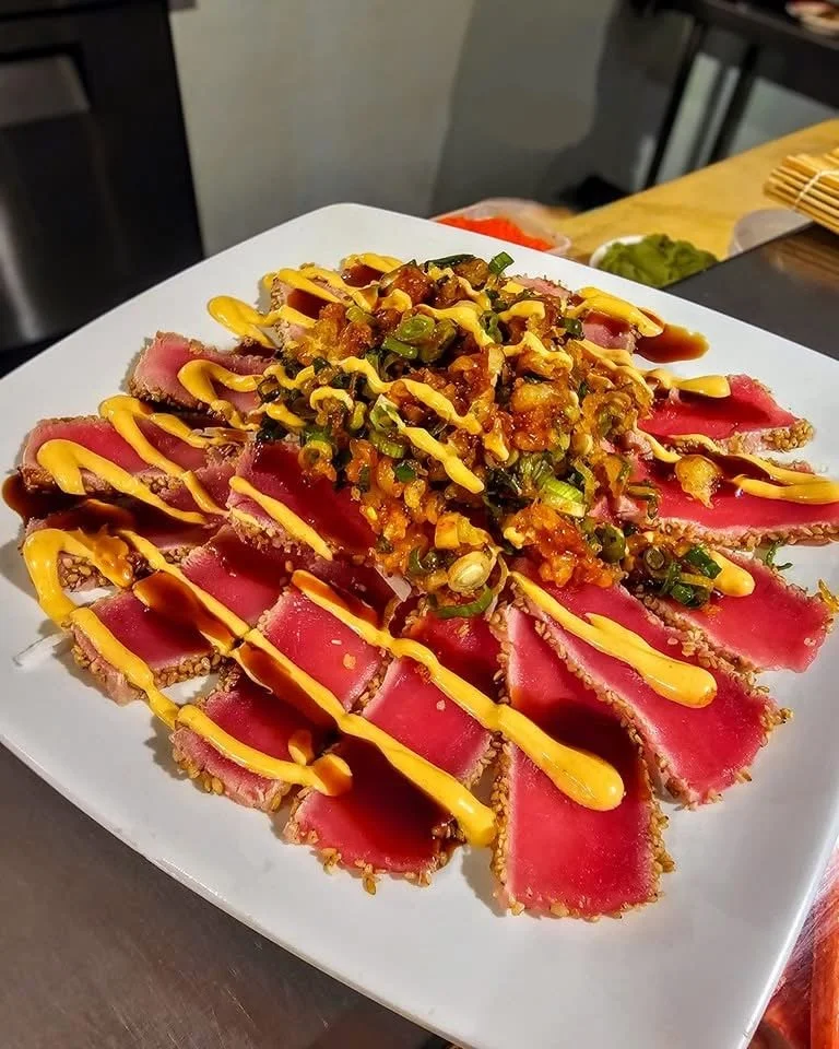 Sliced seared tuna topped with chopped green onions and sauce, garnished with mustard and soy sauce, presented on a white square plate.