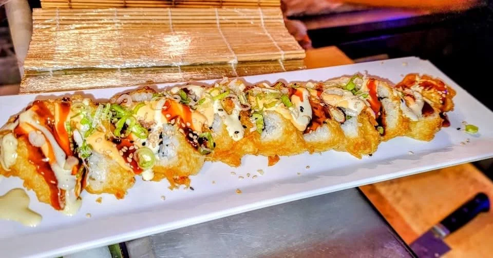 A long white plate with a row of deep-fried sushi rolls topped with sauce, green onions, and sesame seeds.