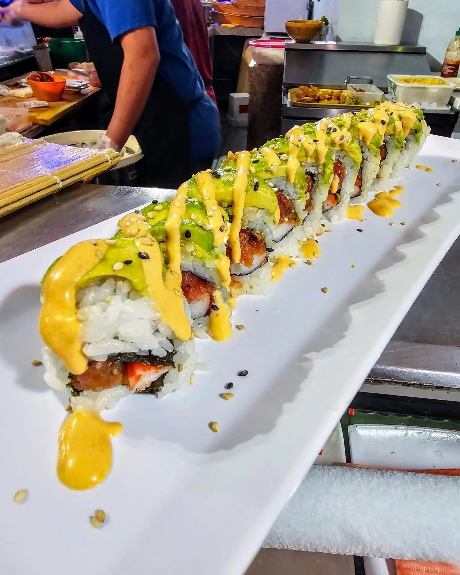 Sushi roll topped with avocado, spicy mayo, and black sesame seeds on a long white plate at a restaurant.