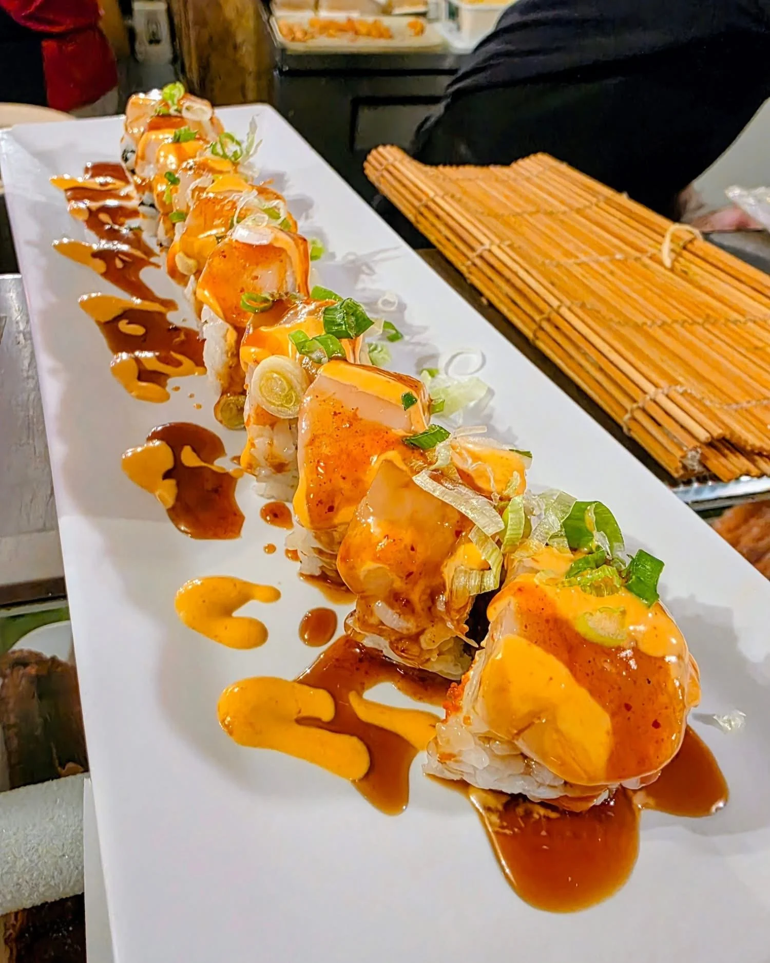 A long white platter of sushi rolls topped with orange sauce, green onions, and drizzled with brown and yellow sauces.