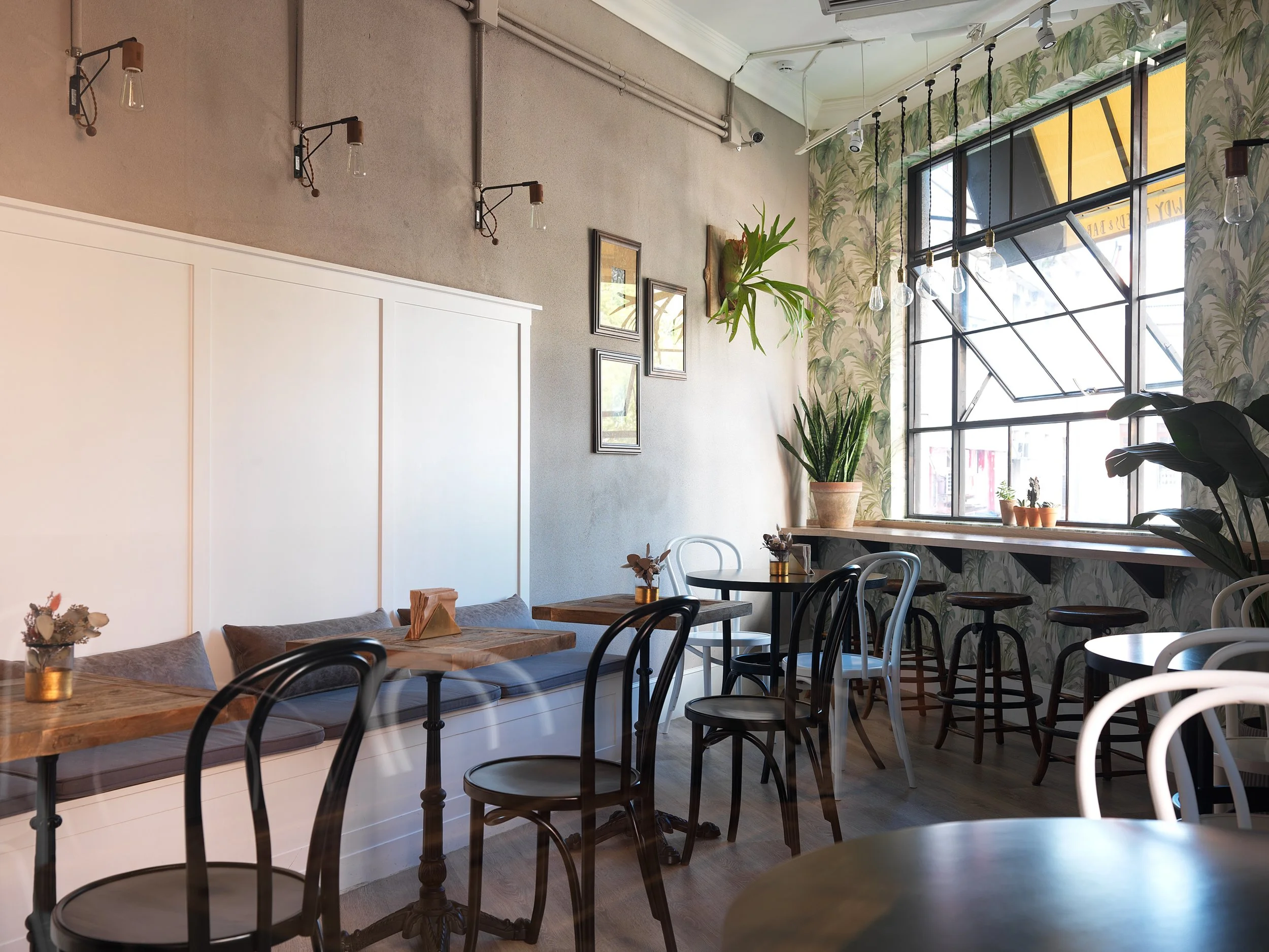 Interior of a cozy cafe with wooden tables and black and white chairs, decorated with potted plants, framed pictures, and large windows with floral curtains letting in natural light.