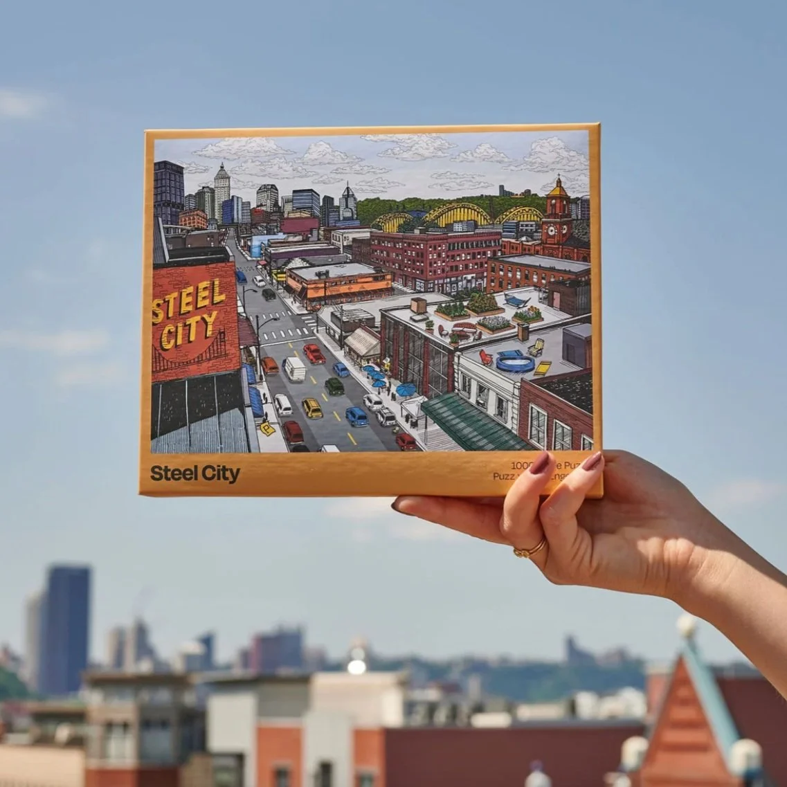 Hand holding a puzzle box titled 'Steel City' depicting a colorful cityscape with buildings, streets, and cars against a blue sky.