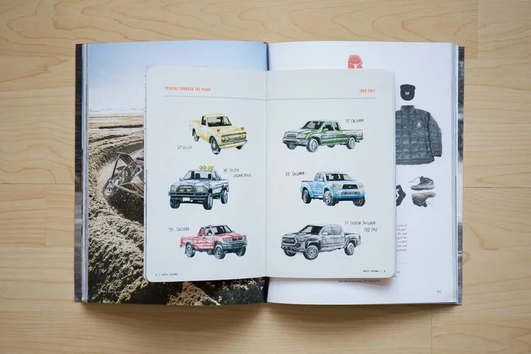 Open magazine displaying illustrated vintage Toyota trucks in various colors on a two-page spread, with a dirt bike and gloves visible on the adjacent page.