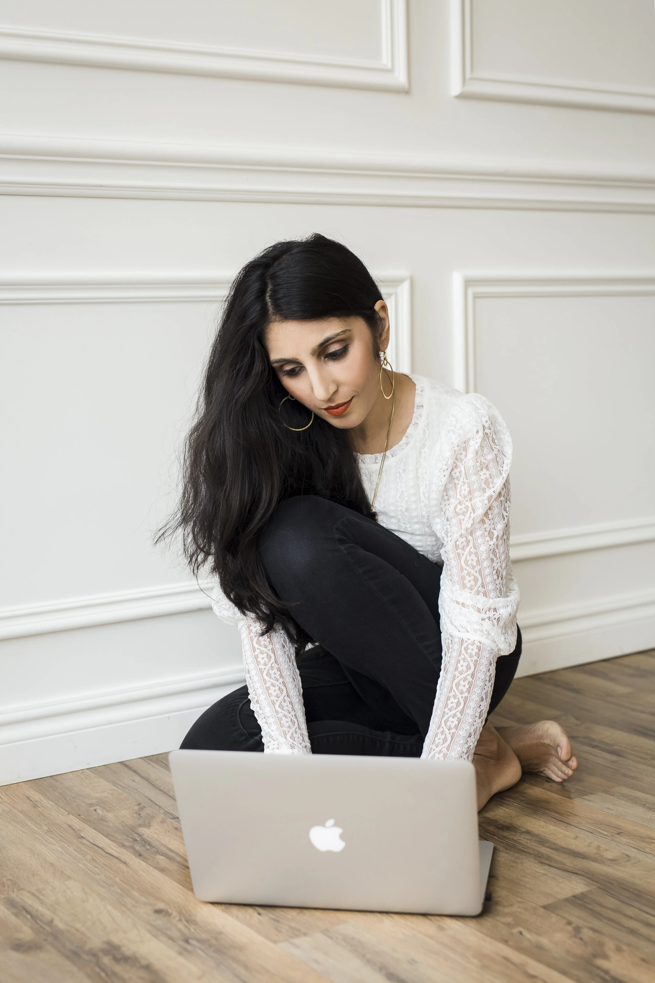 A woman with long black hair, wearing a white lace top, sitting on the floor with her knees held close to her chest and facing a silver laptop, which is open in front of her on a wooden floor. The background features cream-colored wall paneling.