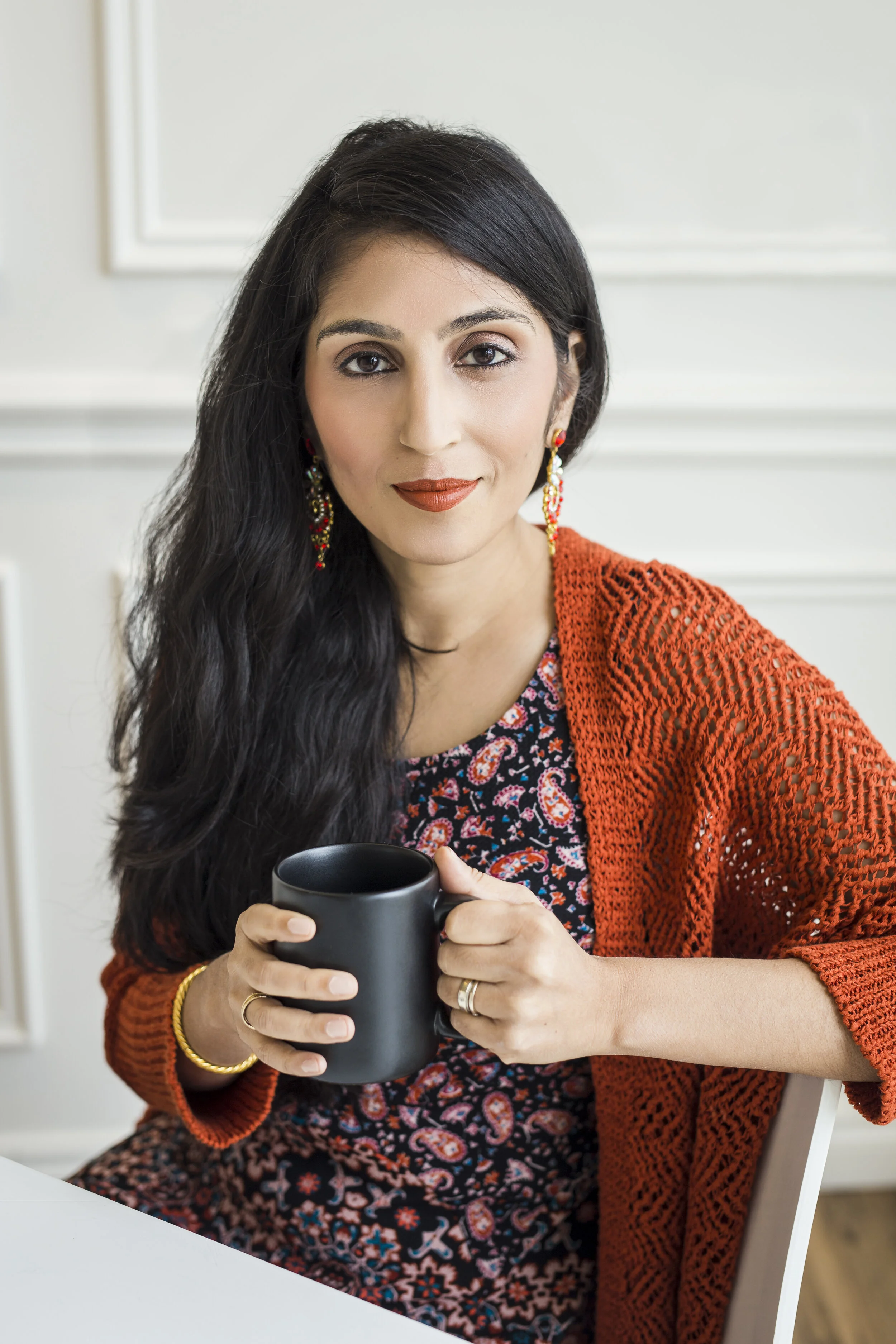 A woman with dark hair wearing a red cardigan and floral dress, holding a black coffee mug, sitting at a white table in a bright room.