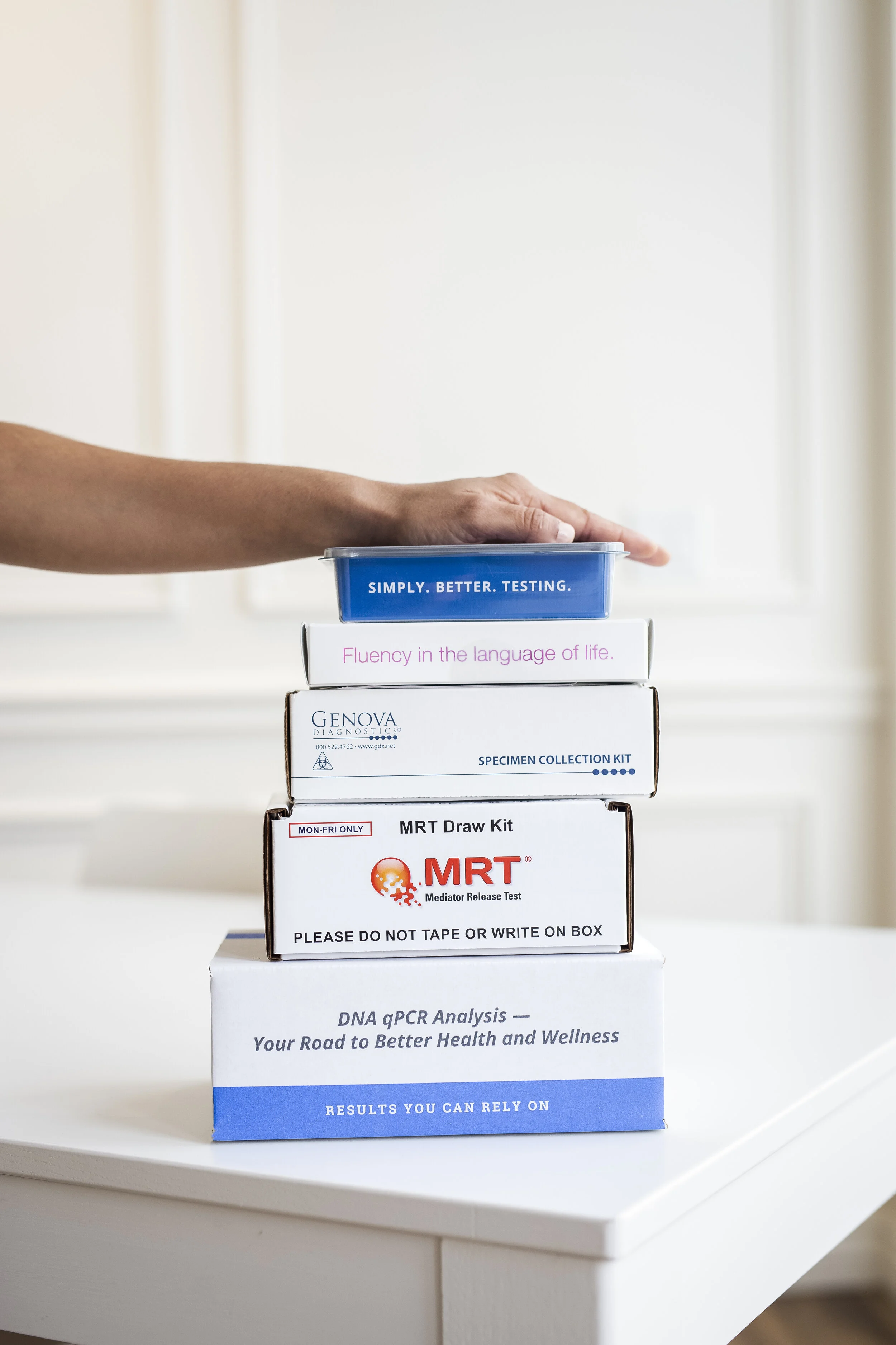 Stacked boxes labeled with various diagnostic and testing kits, including DNA PCR analysis and MRT mediator release test, on a white table.