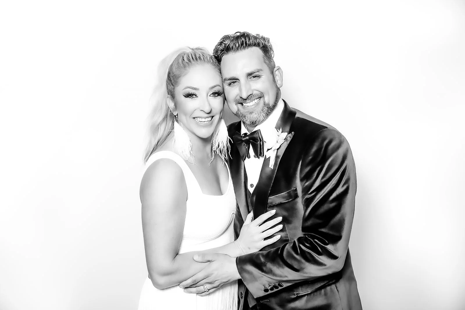 Black and white photo of a smiling couple dressed in wedding attire, standing closely together, with the woman wearing a sleeveless dress and the man in a tuxedo with a bow tie, against a plain background.