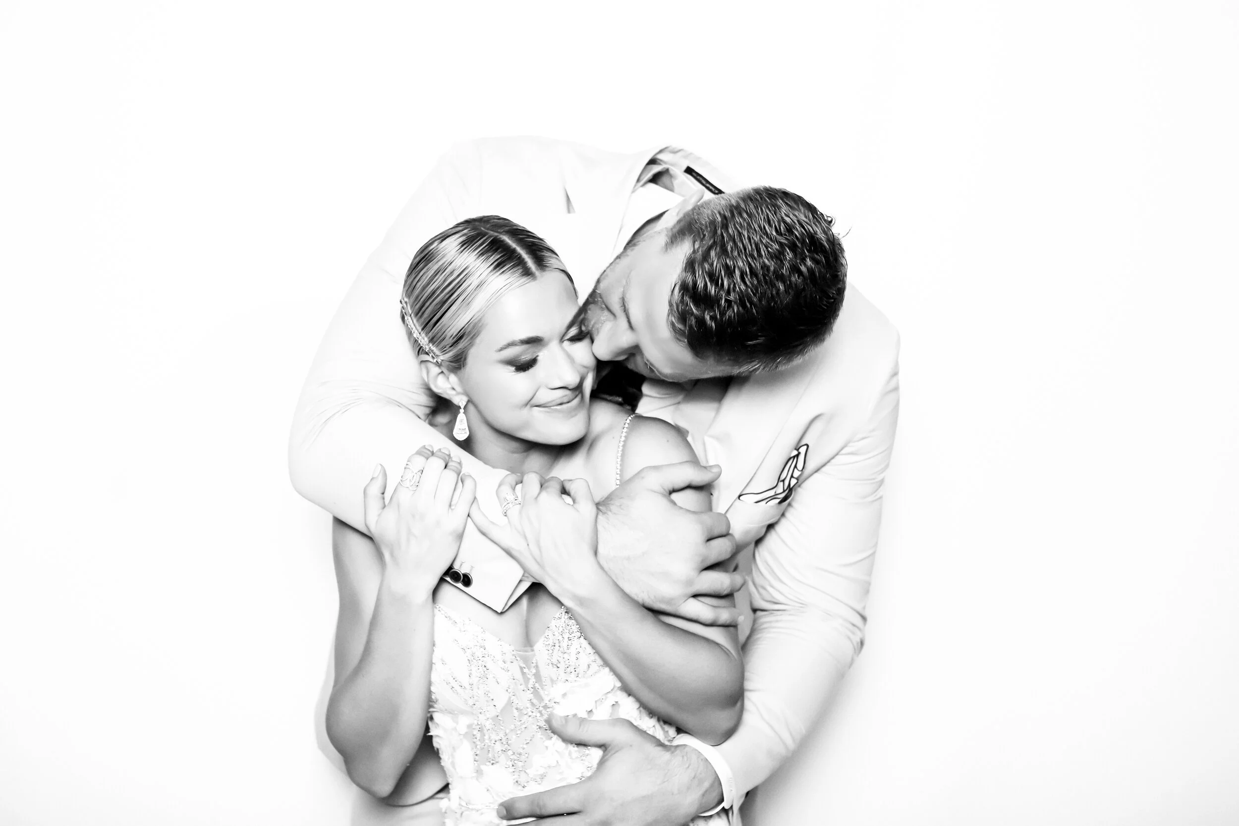 A black and white photo of a man and woman in an embrace, smiling with their eyes closed, against a plain white background.