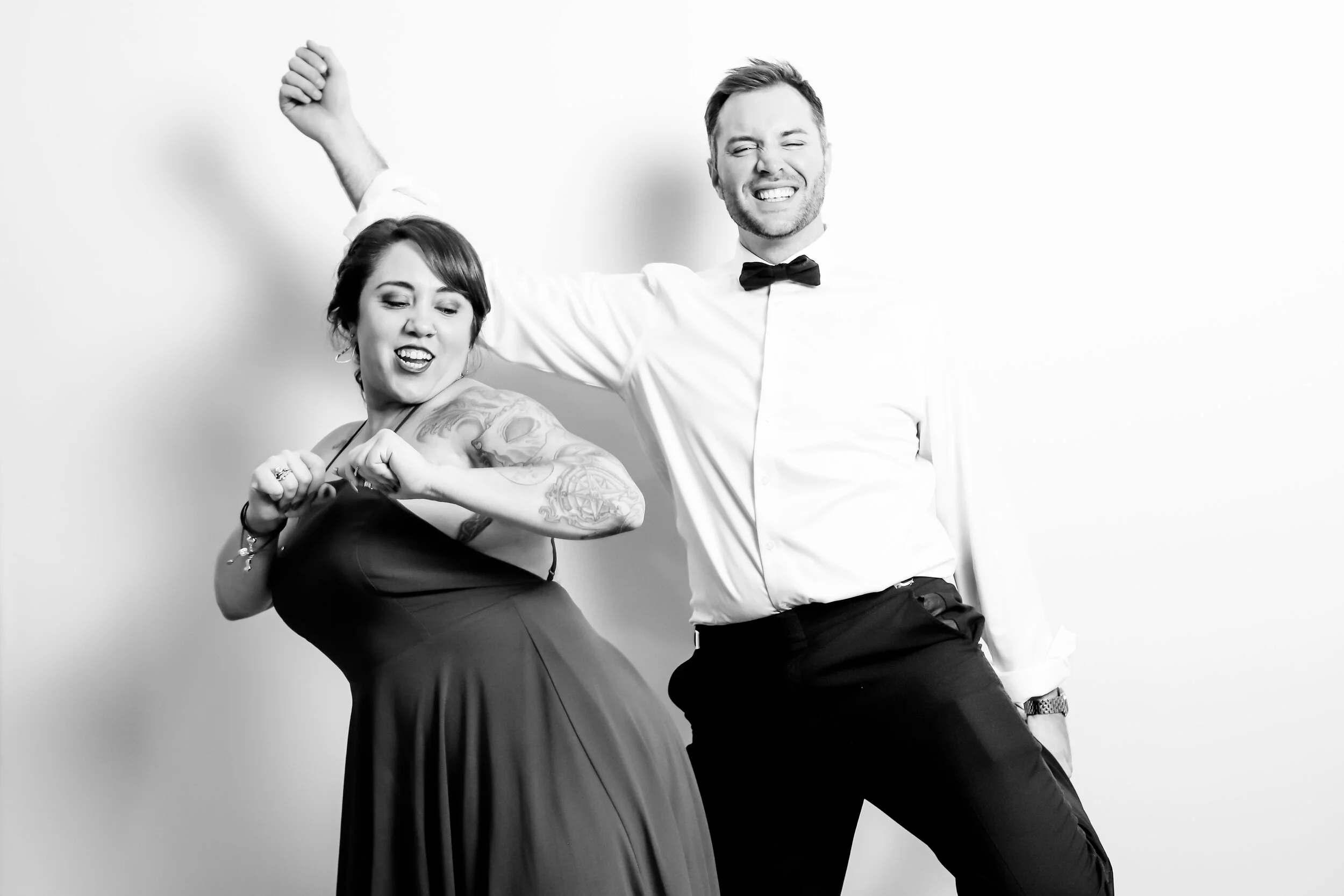 A black-and-white photo of a smiling woman with tattoos and a man in a tuxedo, both dancing or posing energetically against a plain background.