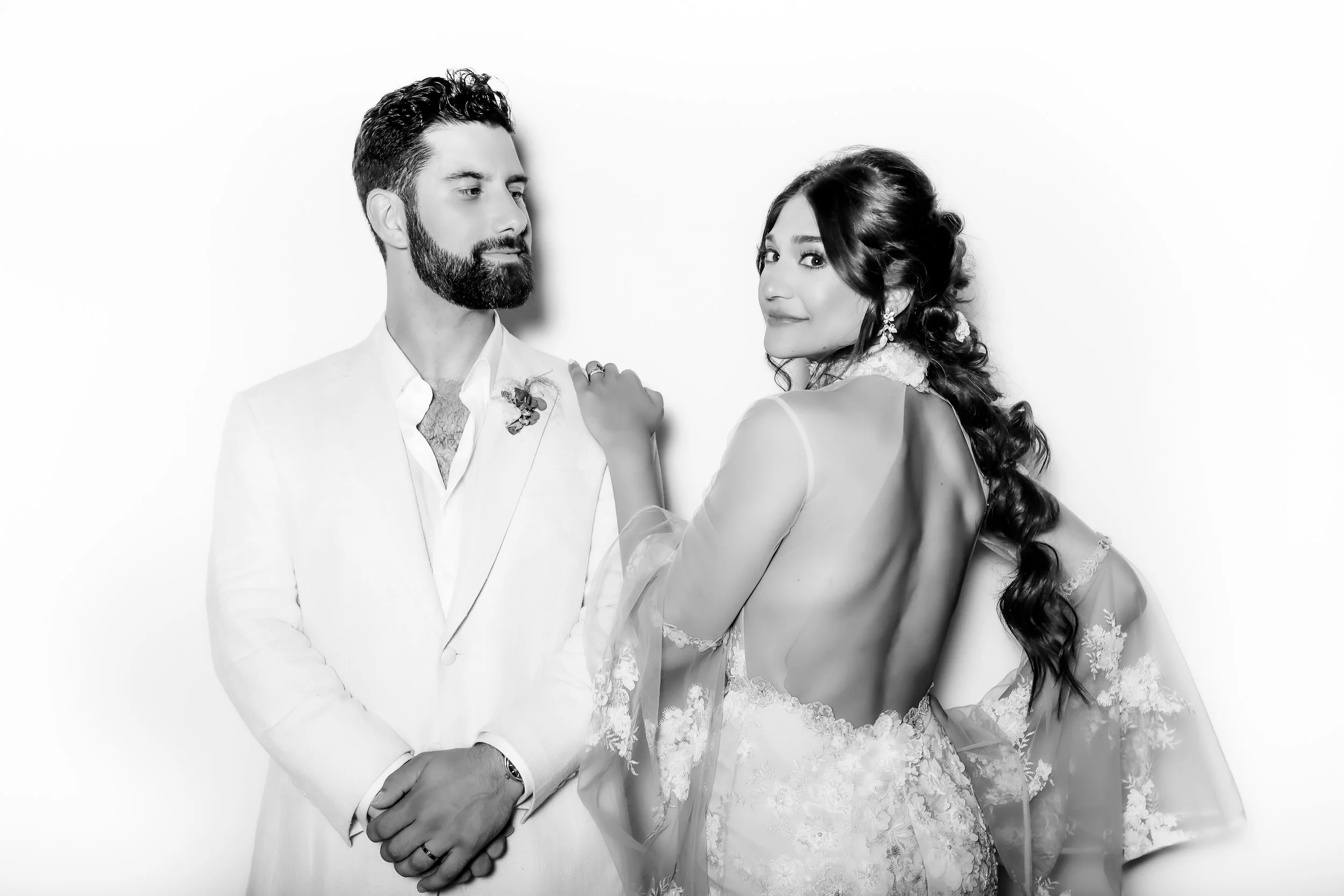 Black and white photograph of a bride and groom, with the groom in a white suit and the bride in a lace wedding dress, standing against a plain white background.