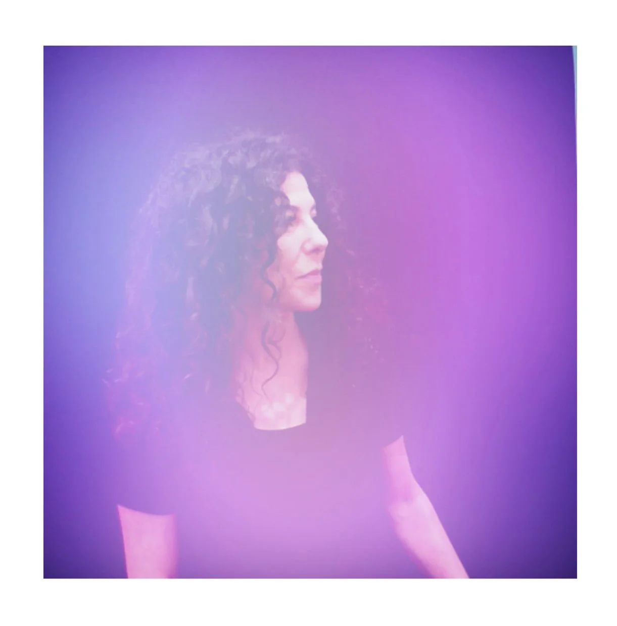 A woman with curly hair dressed in black, looking to the right, with purple and pink lighting creating a soft, dreamy effect.