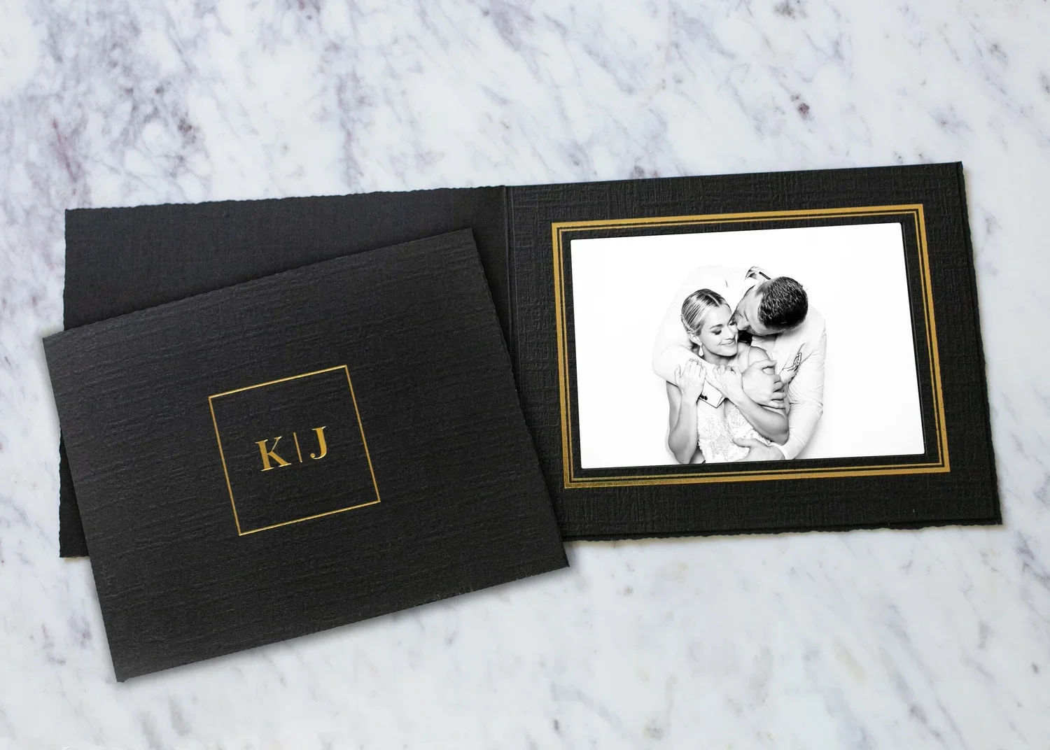 Black photo album with gold initials 'K J' on the cover, opened to show a black and white photo of a couple embracing, on a white marble surface.