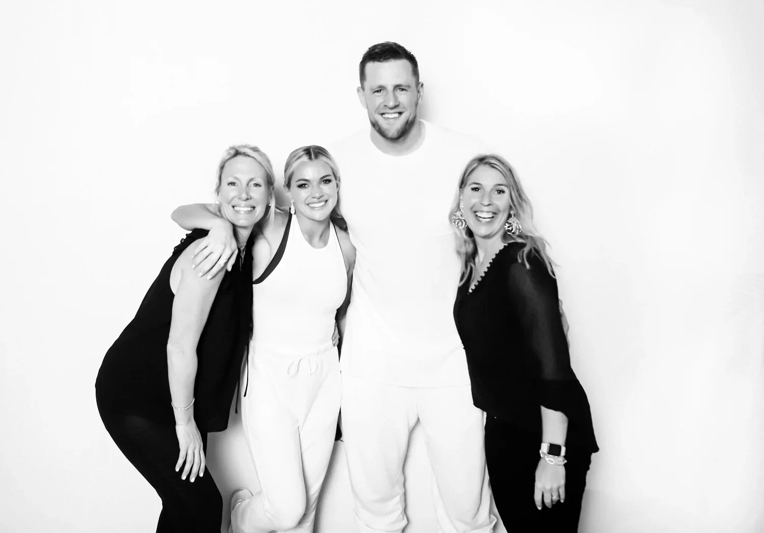 Four people smiling and hugging in front of a plain white wall, with three women and one man, wearing casual and stylish clothing.