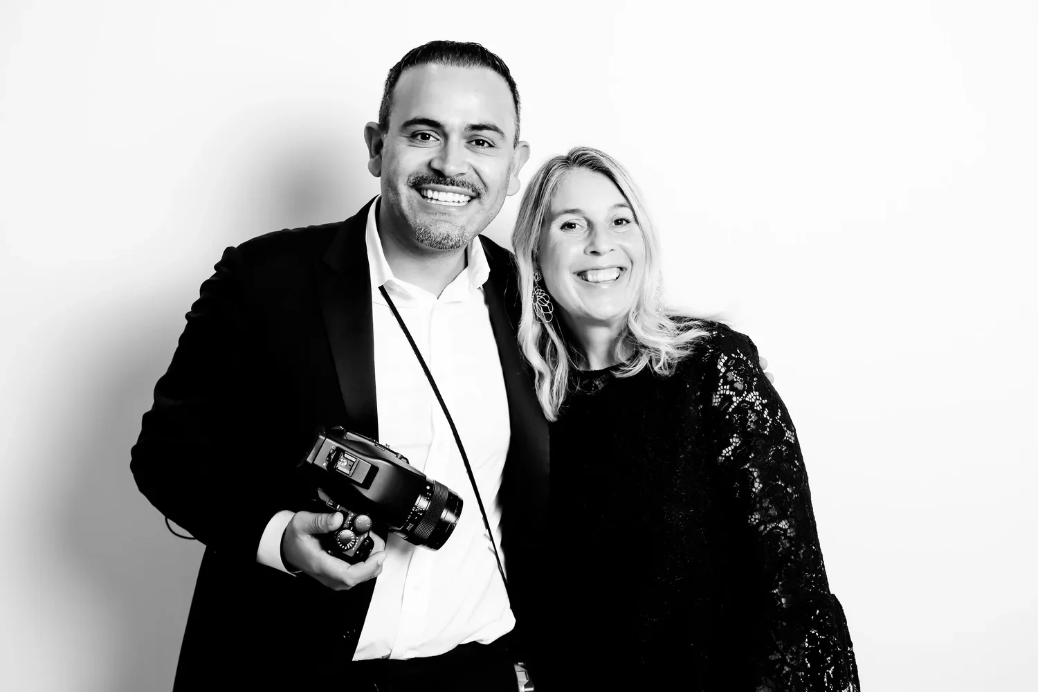 A smiling man in a suit holding a camera stands next to a smiling woman in a lace dress, both posing against a plain background.