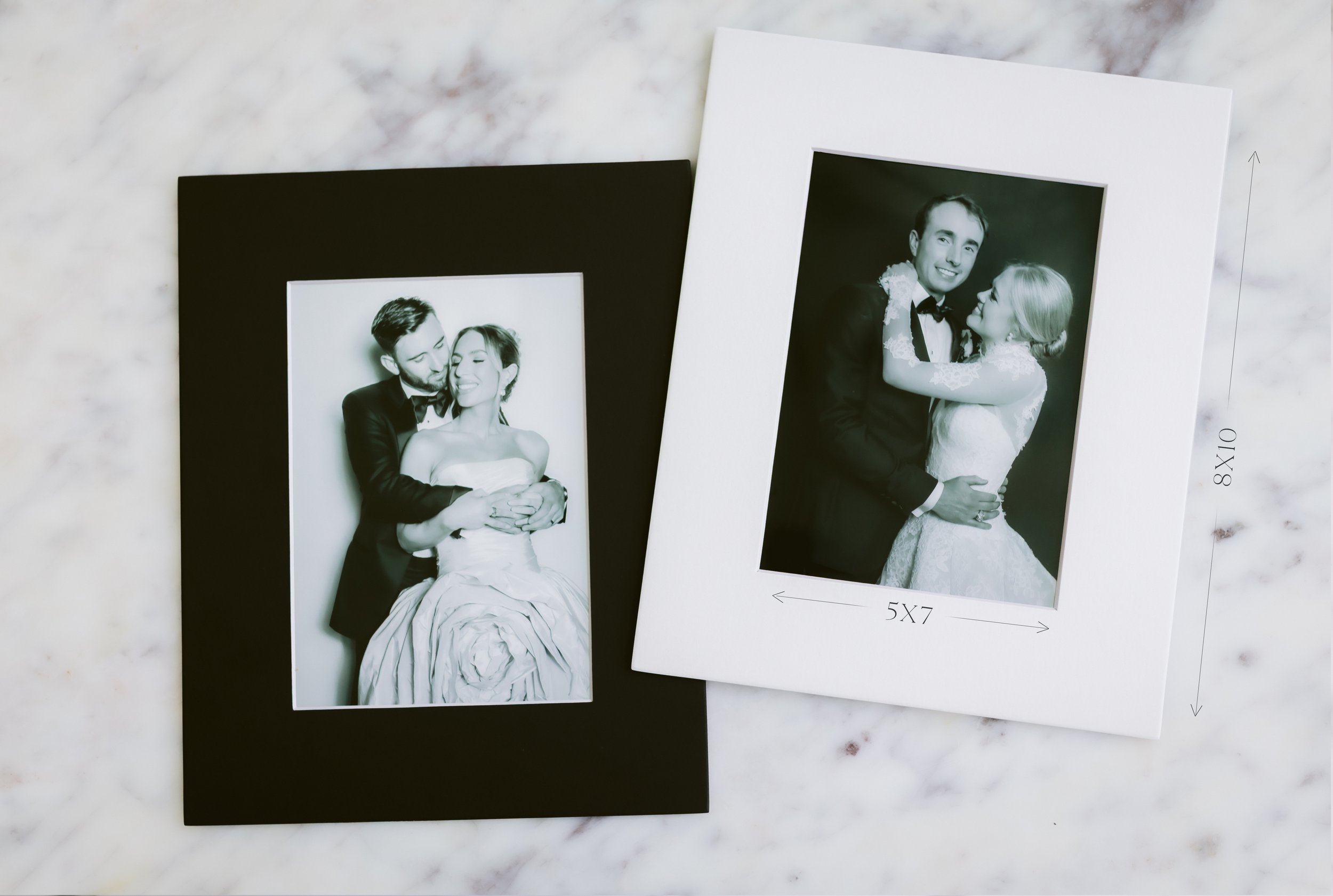 Two printed photographs of a smiling couple in wedding attire, one in black-and-white and the other in color, lying on a white marble surface, with a black background and white frame.