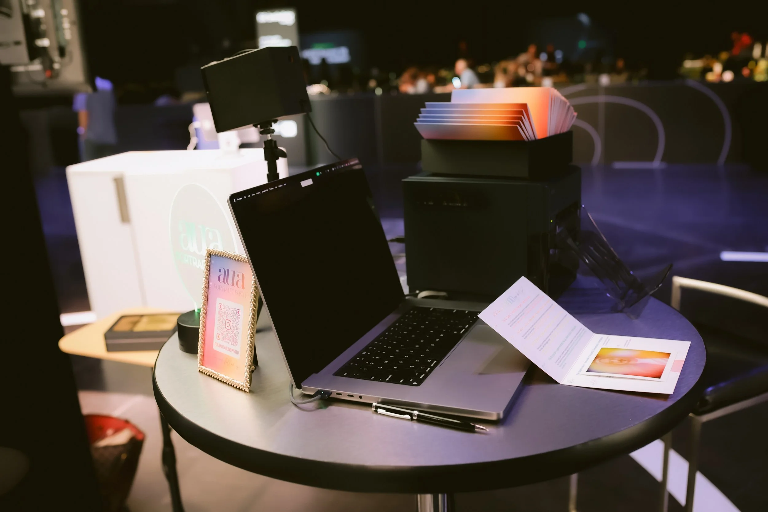 Close-up of the Aura Portrait Booth tabletop setup with laptop capture software, DNP photo printer, and a fanned stack of freshly printed rainbow aura prints