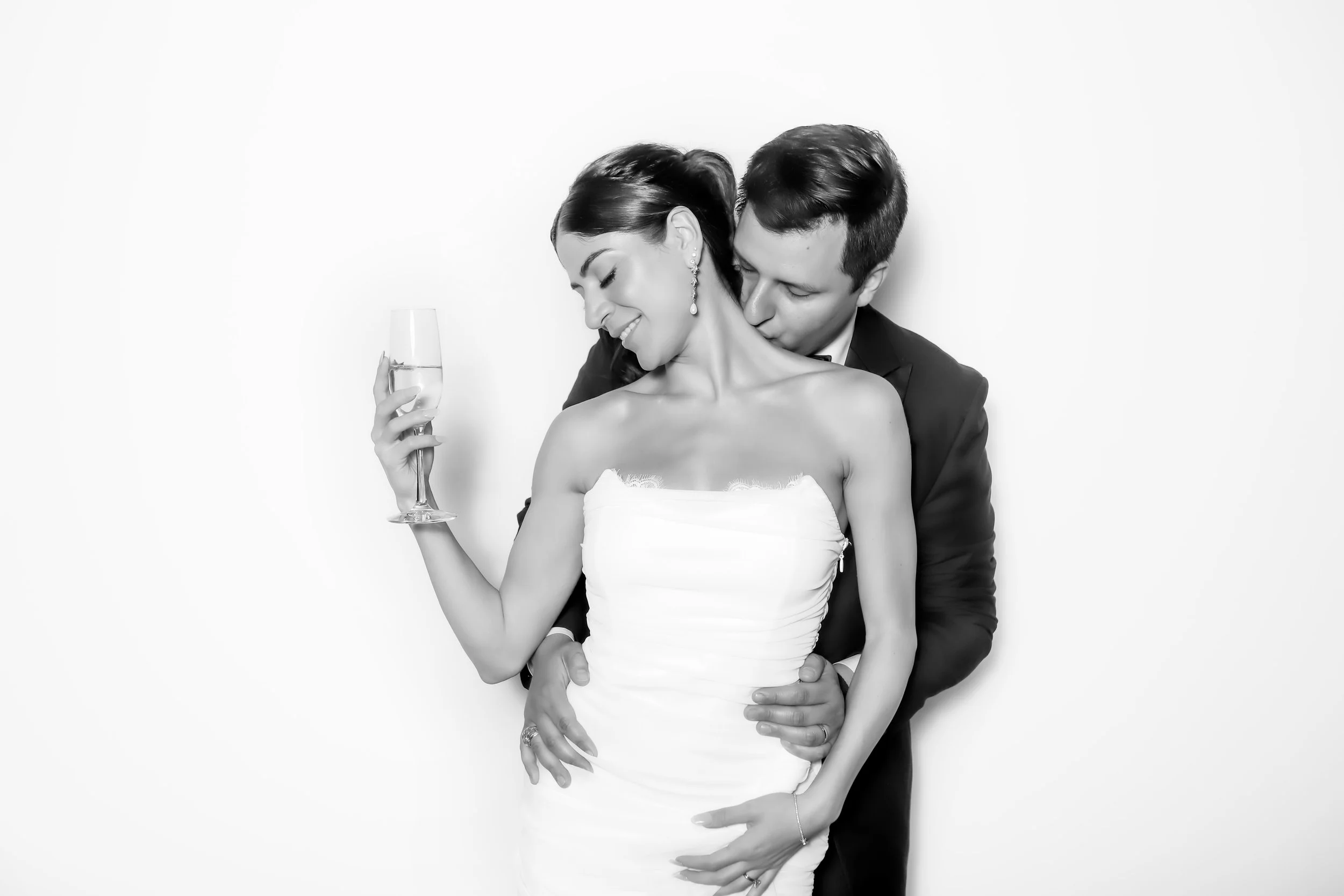 A black-and-white photo of a joyful couple in formal attire, with the woman holding a glass of champagne, the man embracing her from behind, both smiling and looking down, against a plain white background.