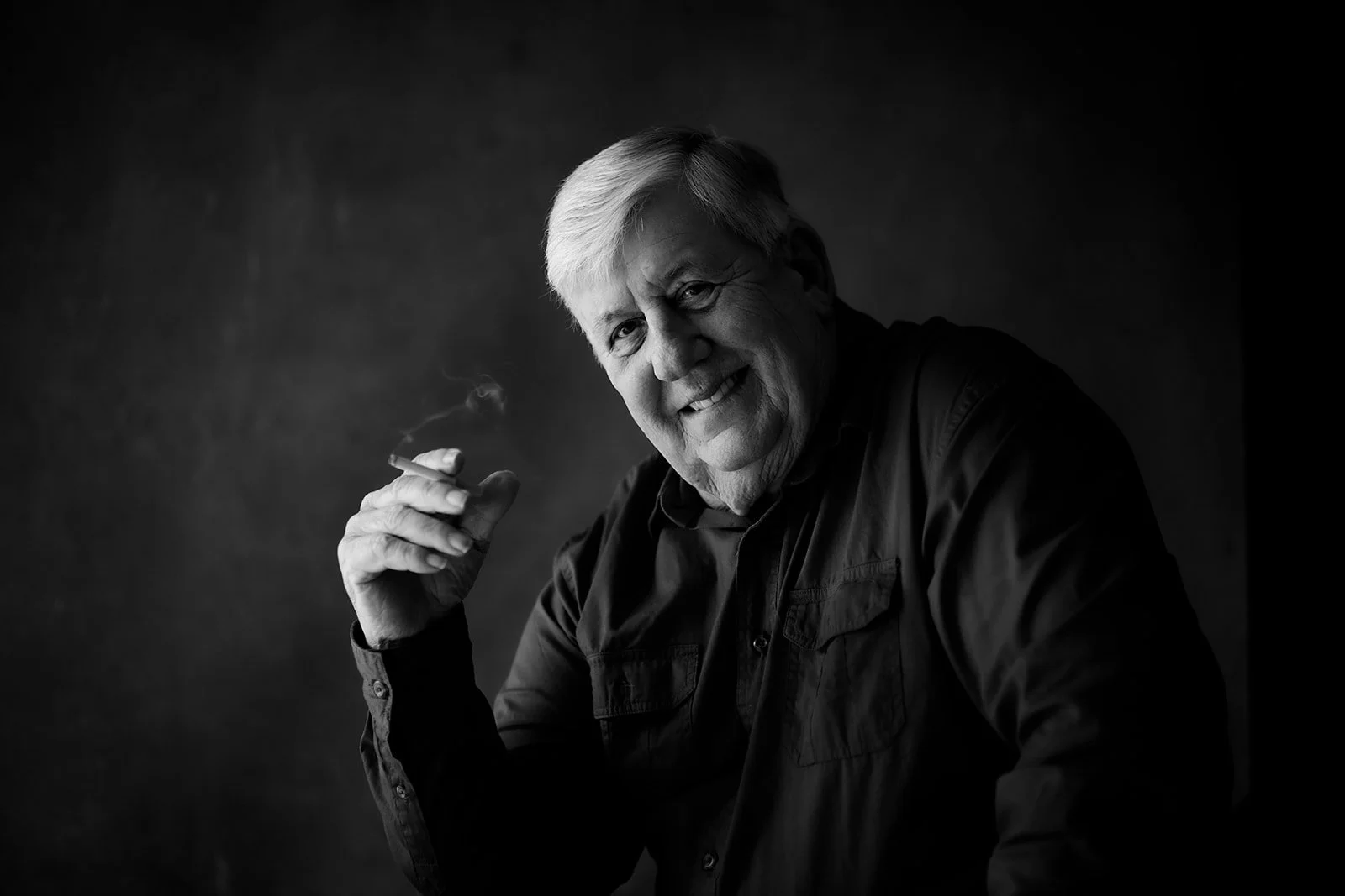 Black and white portrait of an elderly man with light hair smiling and holding a cigarette, against a dark background.