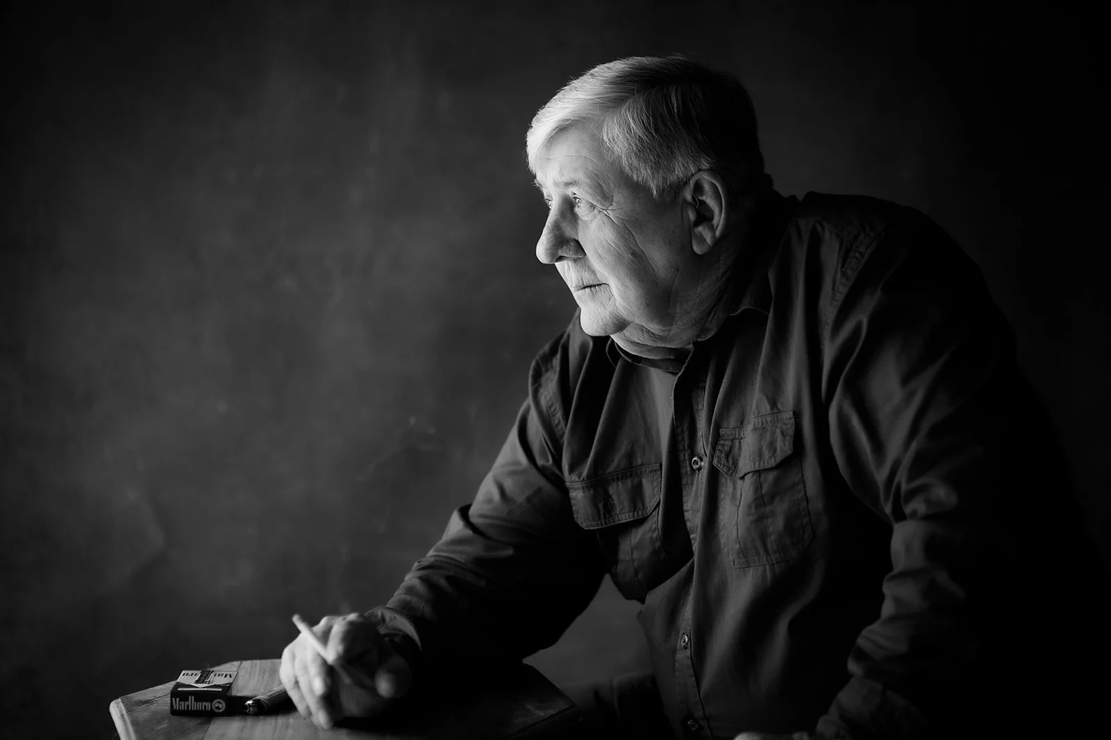 Black and white image of an elderly man with gray hair sitting at a table, looking to his left, with Marlboro cigarette pack and lighter on the table.