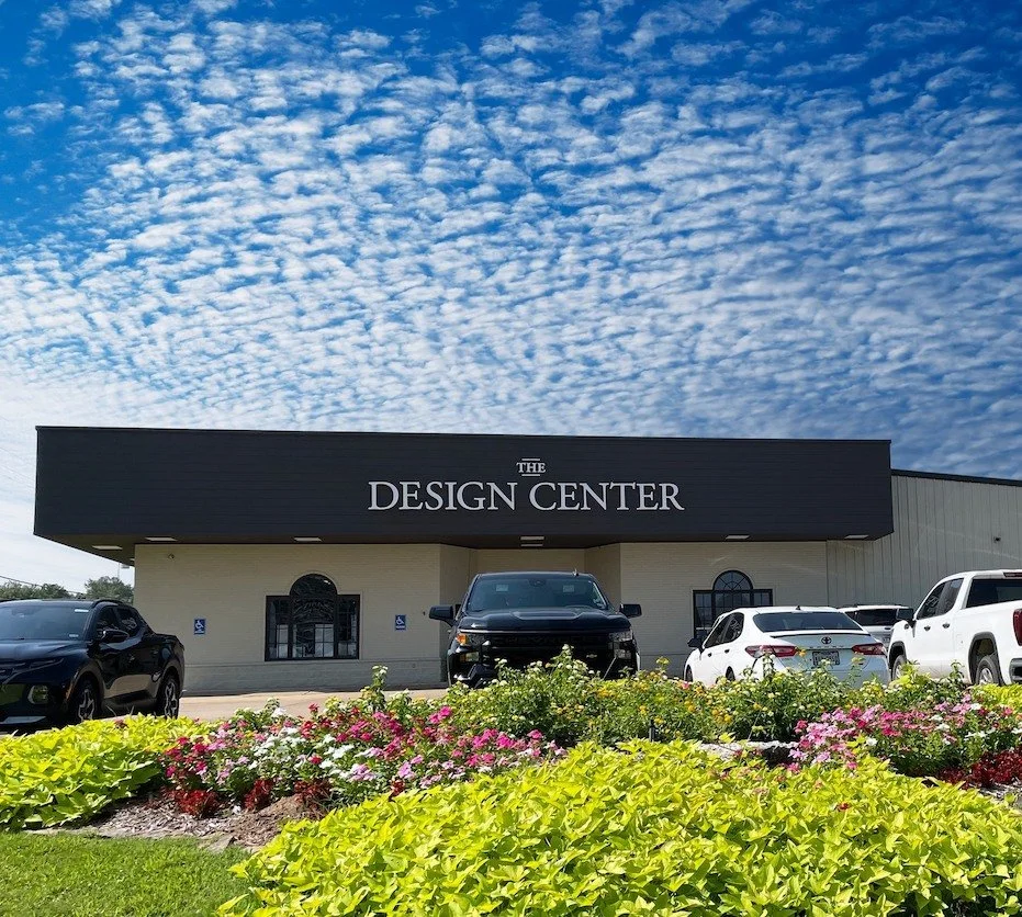 The Design Center building with a black sign, surrounded by a parking lot with cars and colorful flowers, under a blue sky with scattered clouds.