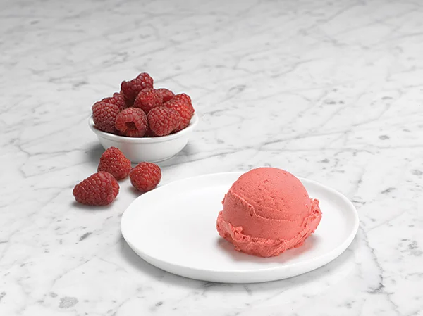 A scoop of pink raspberry ice cream on a white plate with a bowl of fresh raspberries nearby on a marble surface.