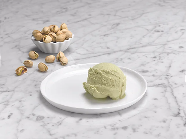 A scoop of pistachio ice cream on a white plate with a bowl of pistachios in the background on a marble surface.