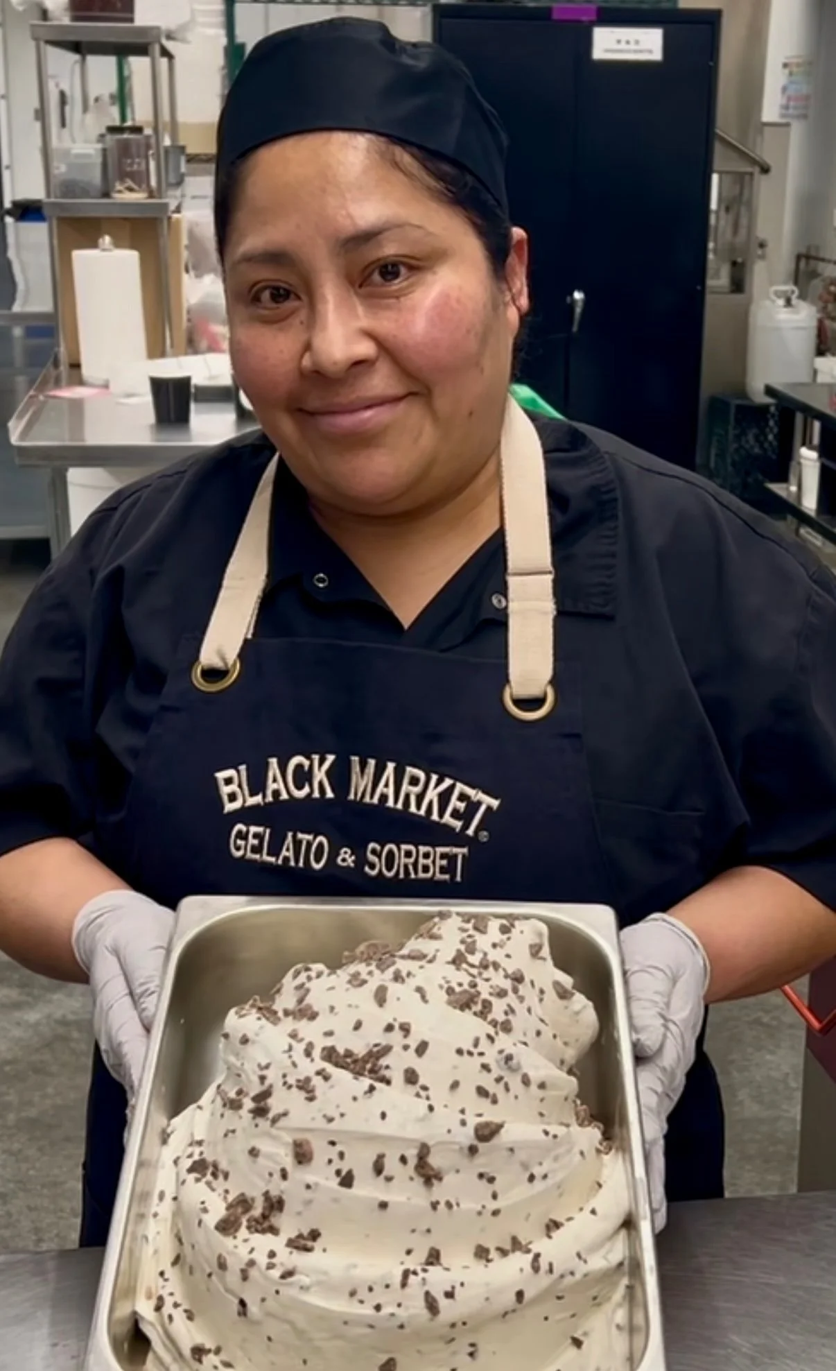 A person in a black apron and cap, holding a metal tray of gelato with chocolate pieces, standing in a commercial kitchen.