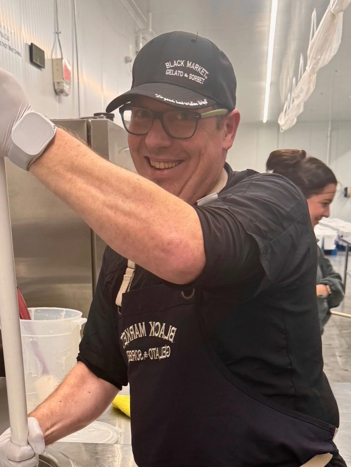 A man smiling at the camera wearing glasses, a black cap with white text, a black apron, and a black shirt, working in a commercial kitchen.