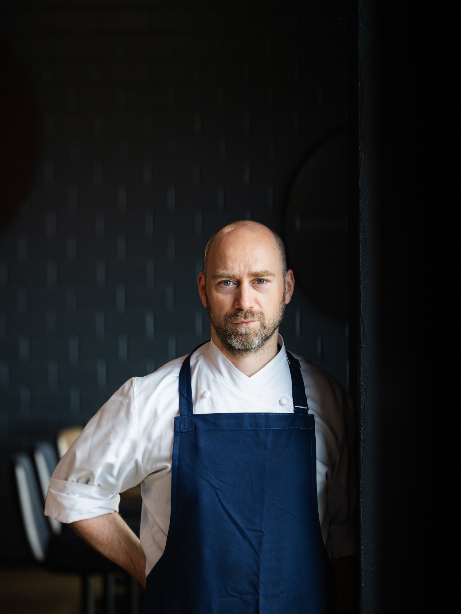 Ein Mann in Kochkleidung, mit blauen Augen, kurzer Bärte, steht in einem dunklen Raum, schaut ernst.