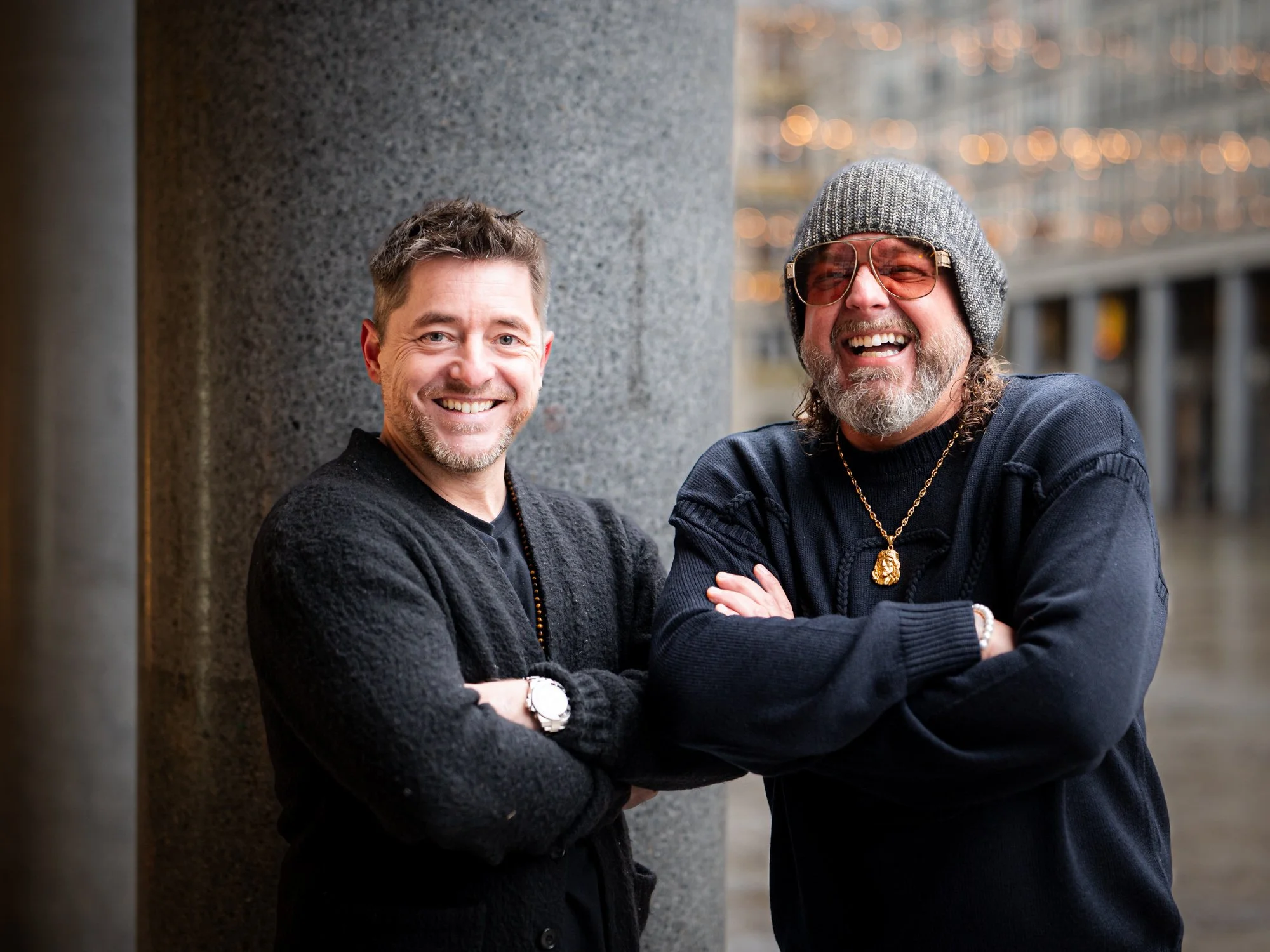 Zwei lachende Männer stehen nebeneinander und haben die Arme verschränkt, in einem urbanen Umfeld, abends mit Lichtern im Hintergrund.