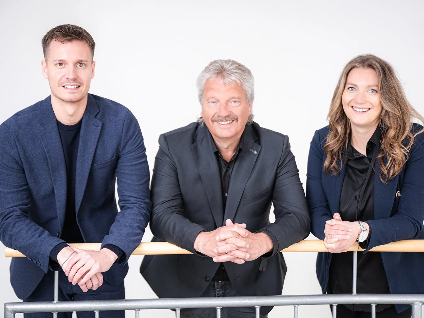 Drei Menschen, zwei Frauen und ein Mann, posieren lächelnd in formeller Kleidung vor weißem Hintergrund.