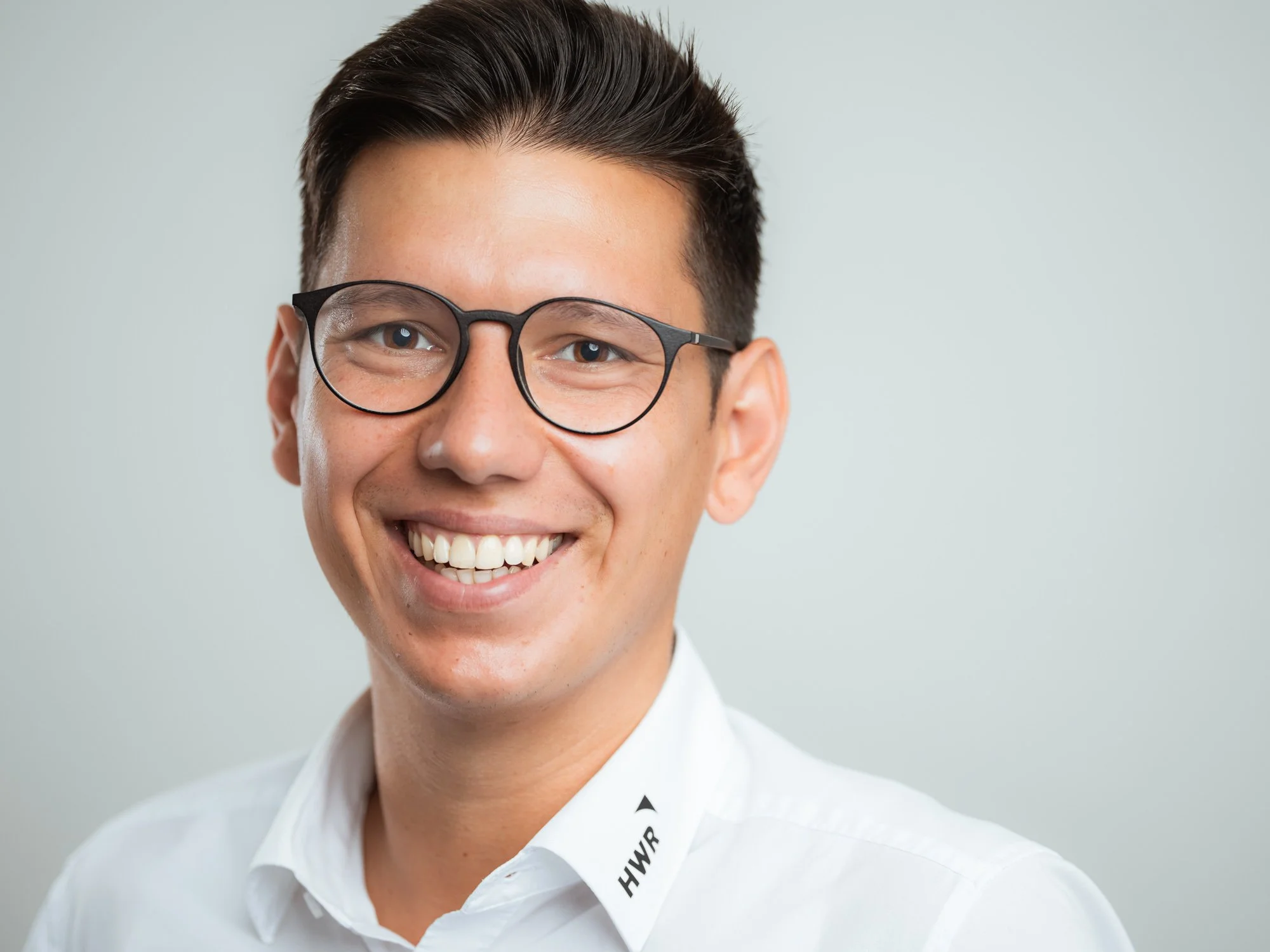 Ein Mann mit dunklen Haaren, Brille und weißem Hemd lächelt vor hellem Hintergrund.