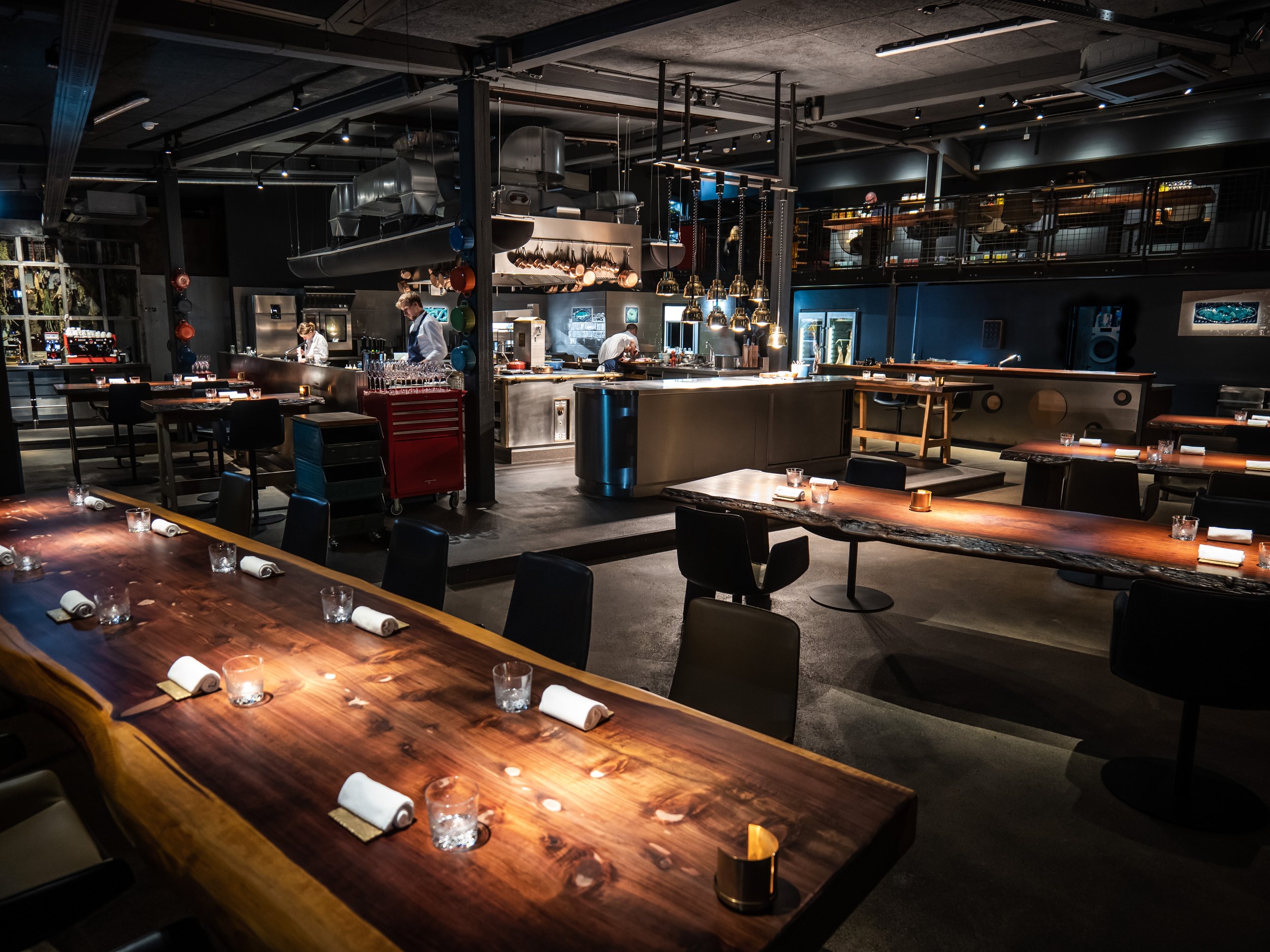 Innenaufnahme eines modernen Restaurants mit offenen Küchenbereich, Holztische, Kerzen und dunkle Stühle, helle Lichtquellen, im Hintergrund Küchenpersonal bei der Arbeit. 100200 Restaurant Hamburg