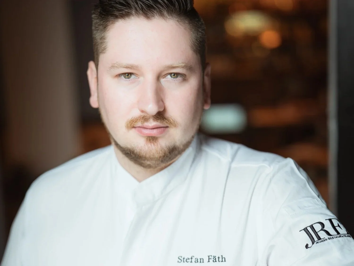 Portrait eines männlichen Kochs in weißer Kochjacke, mit Namen Stefan Fäth, in einem Restaurantküchenhintergrund.