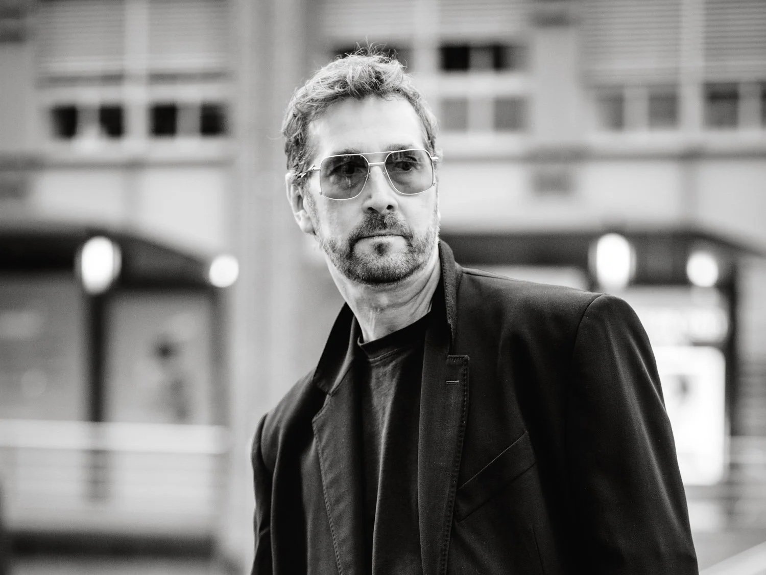 Schwarz-weiß-Foto eines Mannes mit kurzen Haaren, Bart und Sonnenbrille. Er trägt einen dunklen Mantel und einen dunklen Pullover und steht vor einem städtischen Hintergrund mit Gebäuden.