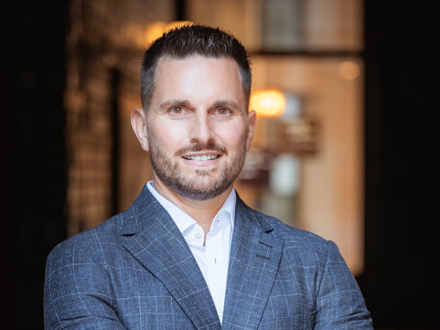 Porträt eines lächelnden Mannes in einem blauen Anzug vor einem warm beleuchteten Hintergrund.