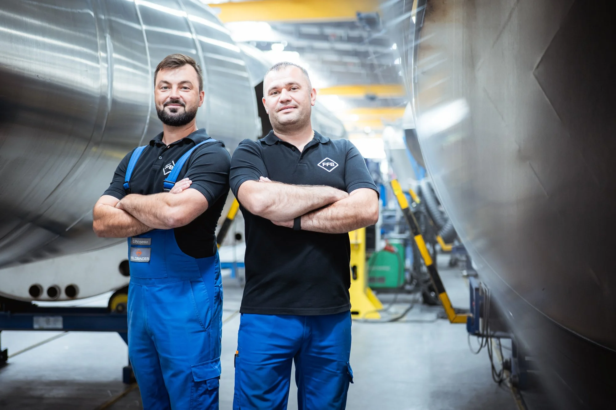 Zwei Männer in Arbeitskleidung stehen mit verschränkten Armen in einer Werkstatt zwischen großen Metallbehältern.