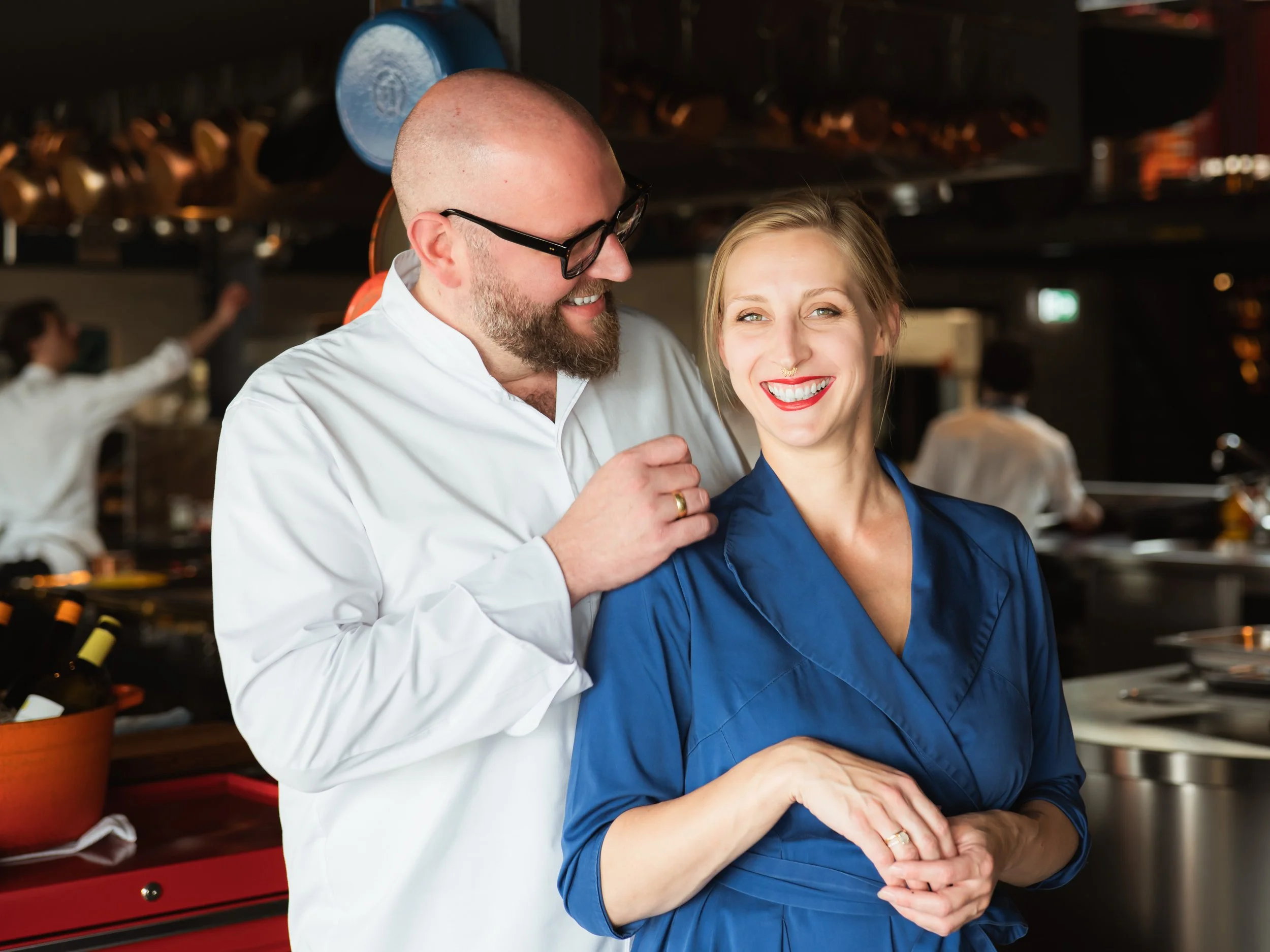 Ein Koch in einer weißen Kochjacke lächelt eine Kellnerin in einer blauen Uniform an, sie lächelt ebenfalls. Sie befinden sich in einer professionellen Küche oder einem Restaurant.