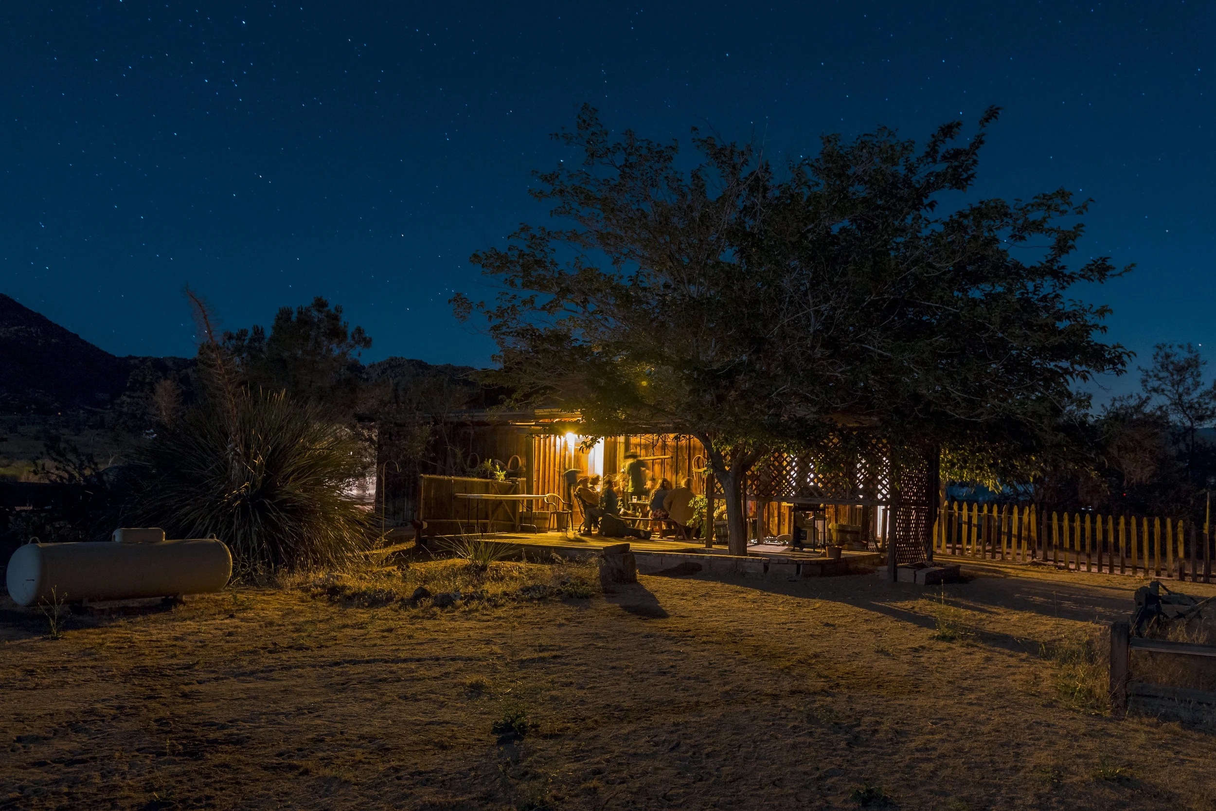 Nighttime scene of a house with warm lighting, people sitting outside under a large tree, stars in the sky, surrounded by mountains and natural landscape.