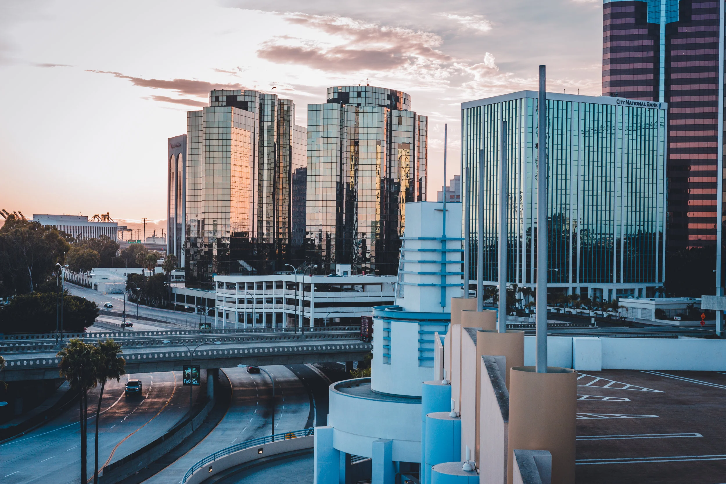 Cityscape featuring modern glass high-rise buildings, a parking garage, and a highway at sunset with a pink and orange sky.