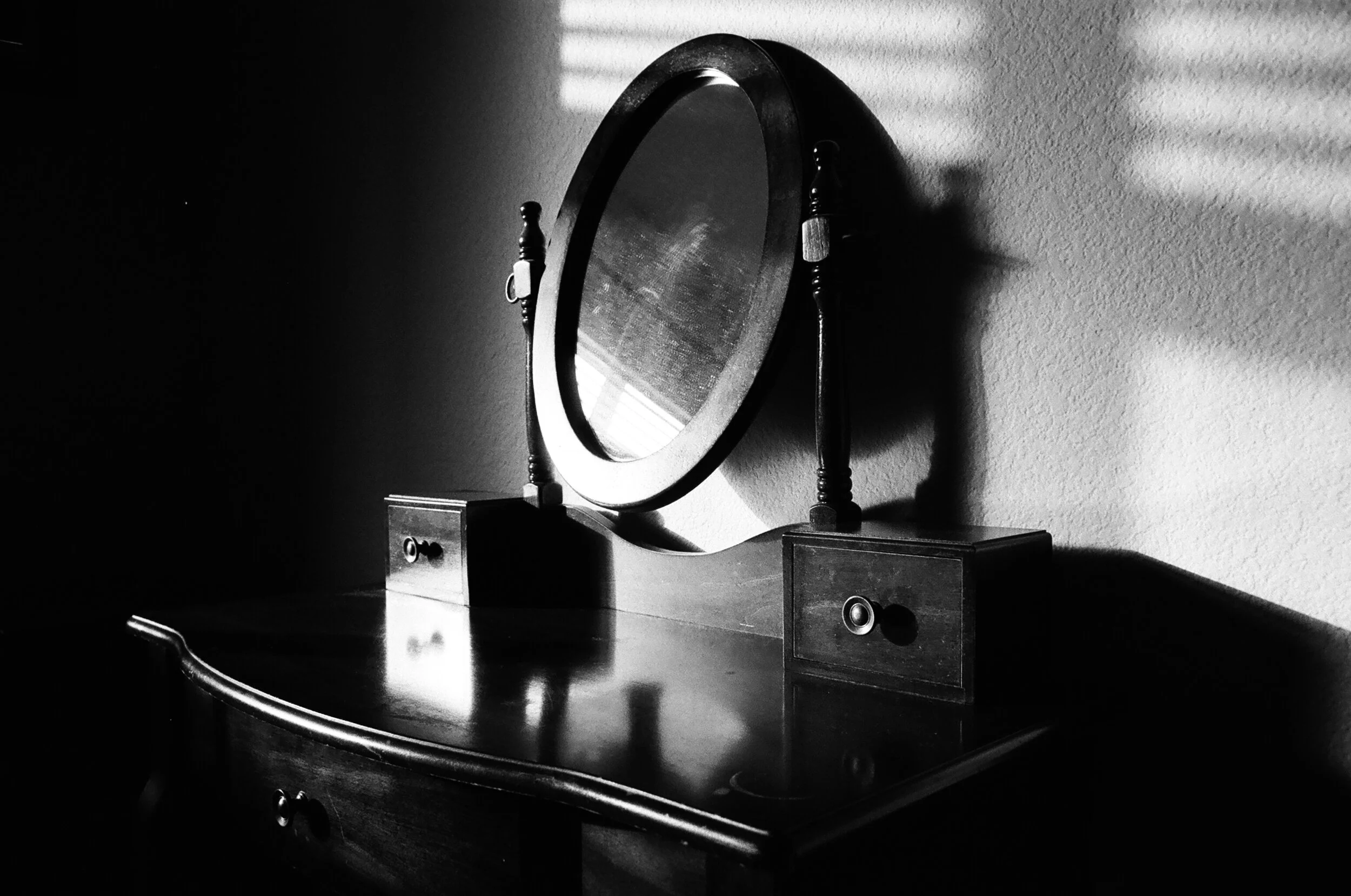 A black and white photograph of an antique wooden dresser with a mirrored top, ornate knobs, and shadows from window blinds cast on the wall.