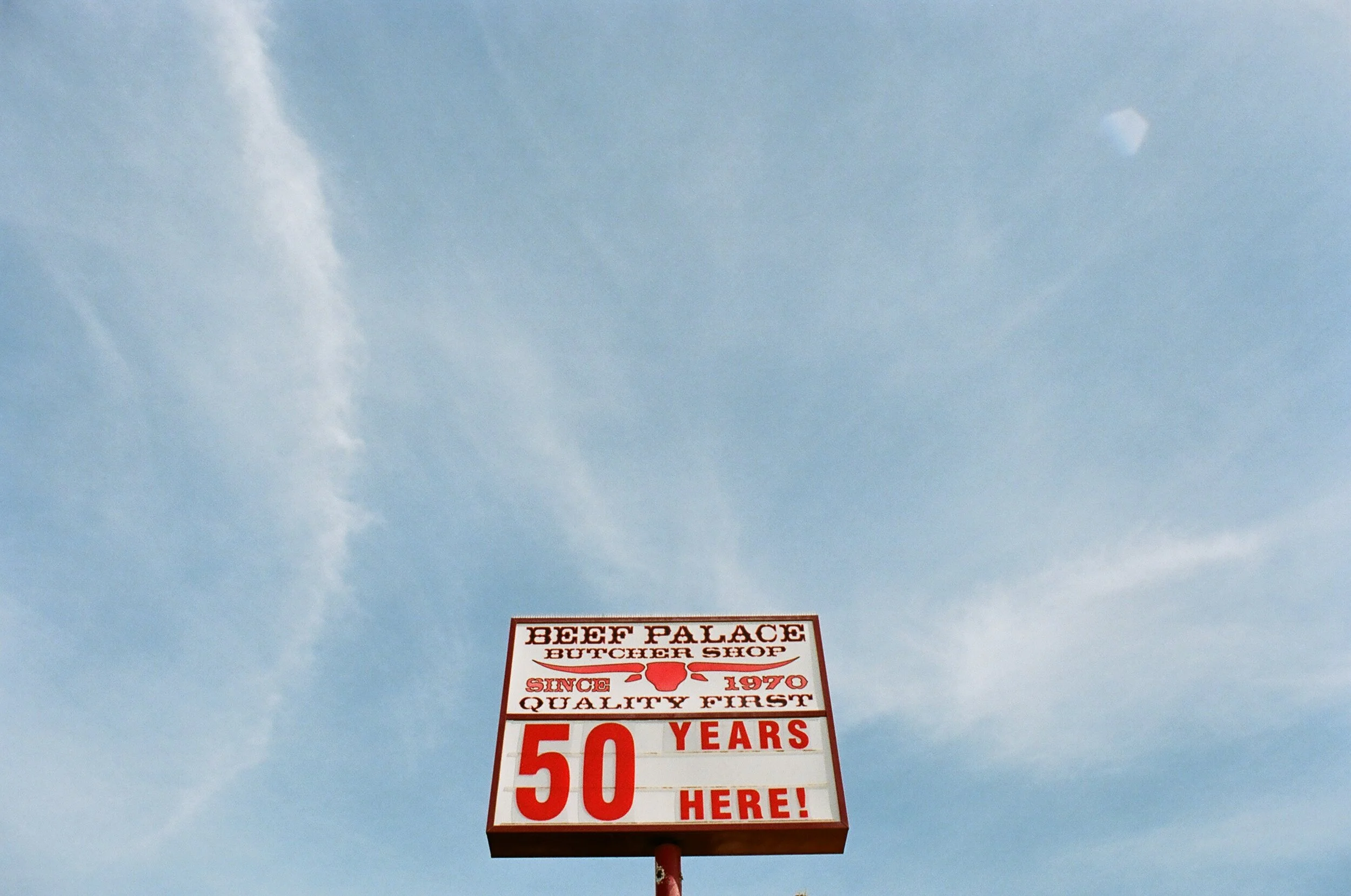 A signboard in the sky reading "BEEF PALACE BUTCHER SHOP SINCE 1970 QUALITY FIRST 50 YEARS HERE!" against a light blue sky with some clouds and a faint visible sun.