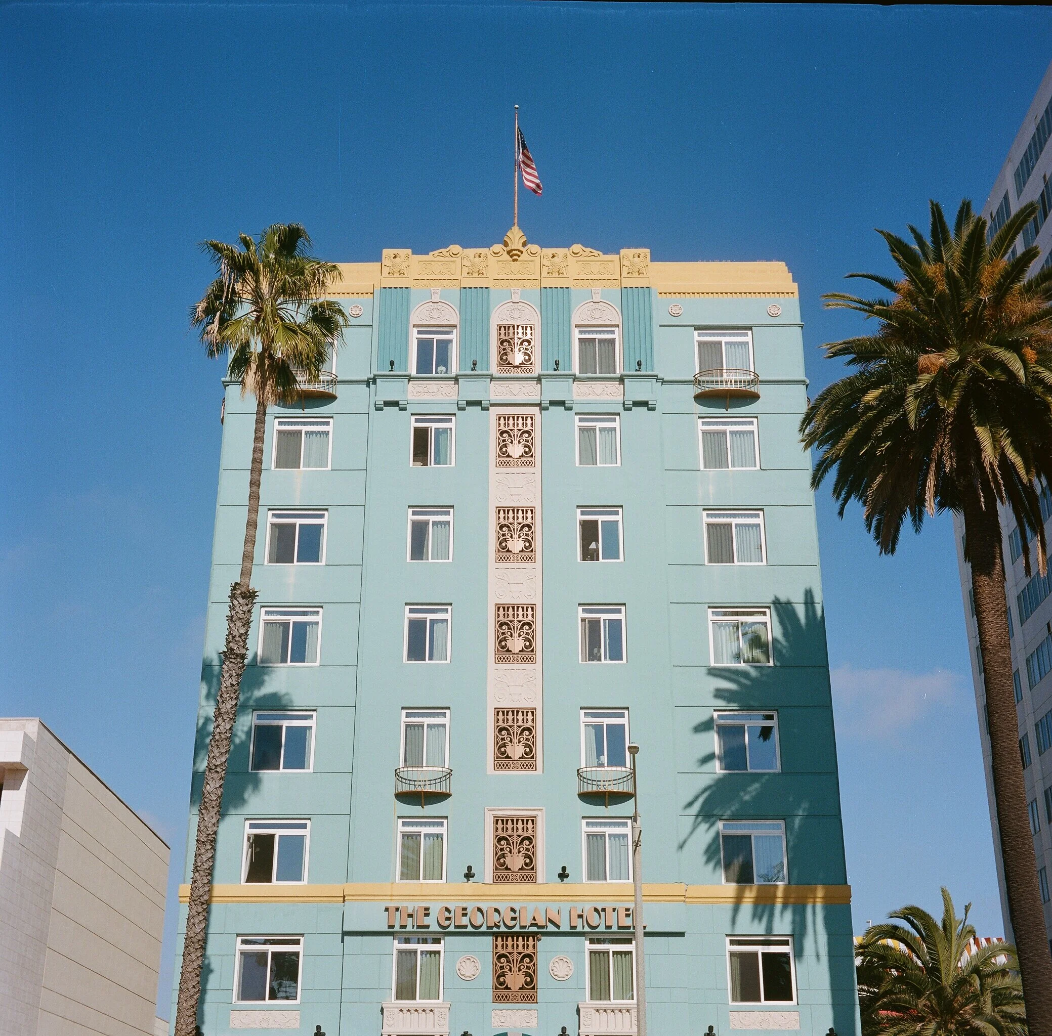 A tall, vintage-style building called The Georgian Hotel with a light blue facade, decorative architectural details, and an American flag on top. There are palm trees casting shadows in front of the building.